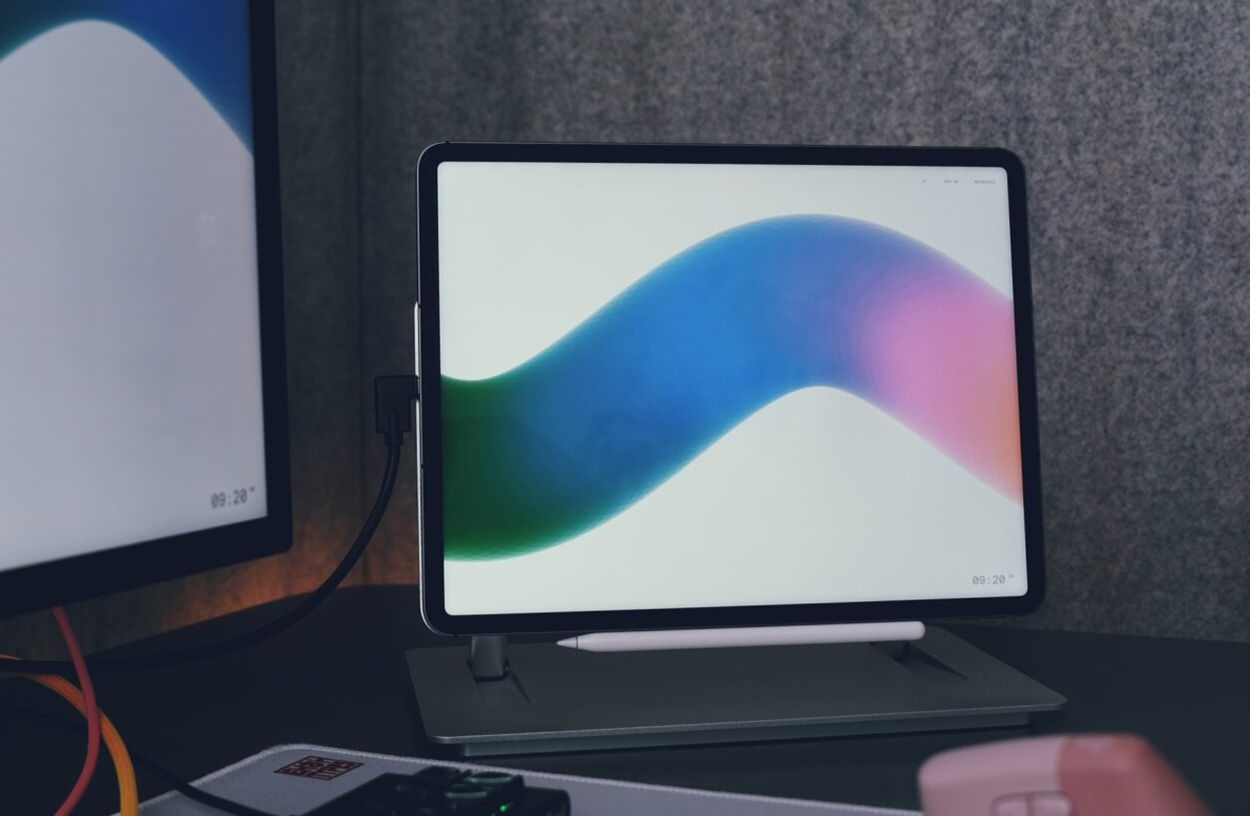 Auf dem Bild sieht man ein Tablet mit einem farbenfrohen Hintergrund, das auf einem Ständer auf einem Schreibtisch steht. Darunter liegt ein Stylus, und im Hintergrund ist ein Teil eines Monitors zu sehen. Die Szene wirkt modern und aufgeräumt.