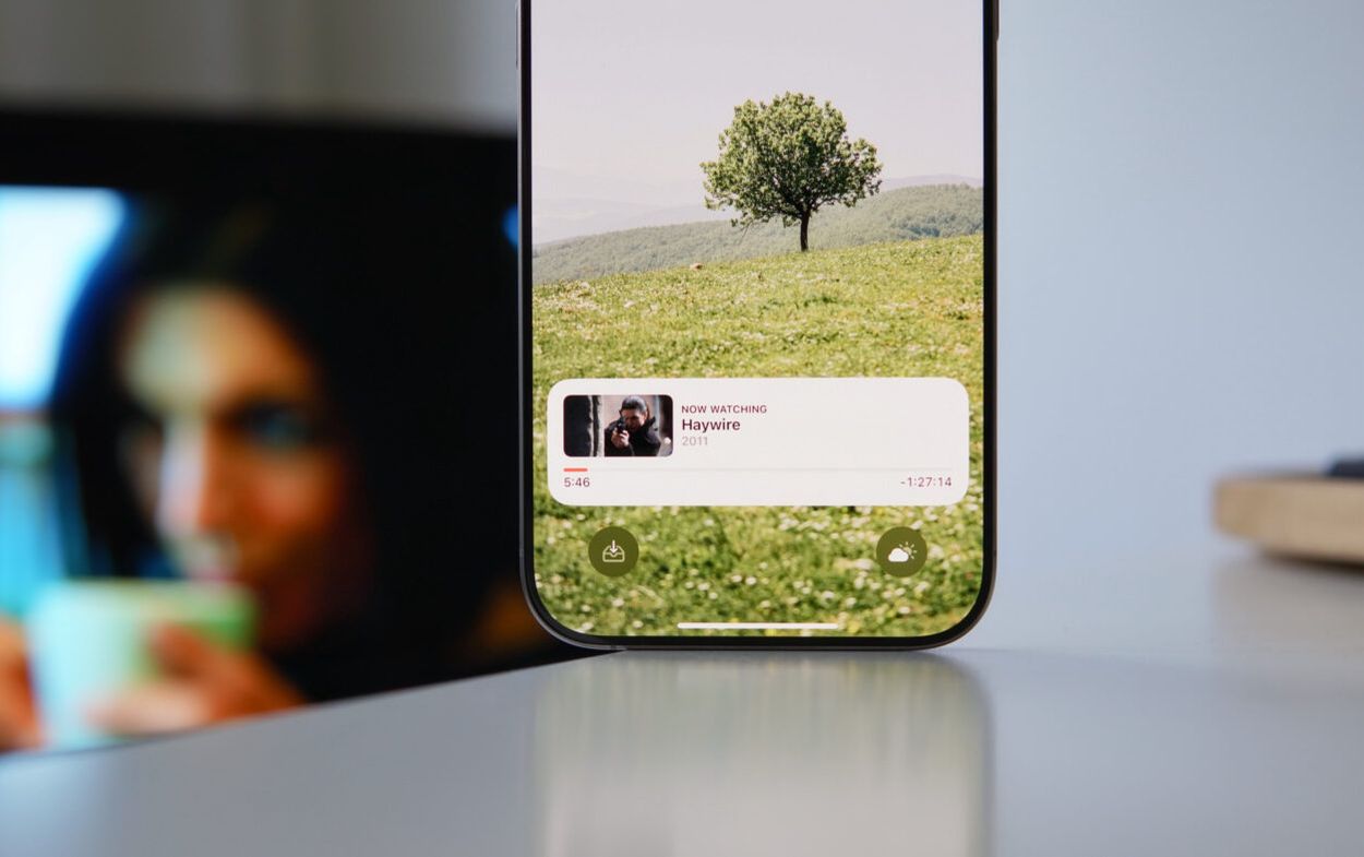 Auf diesem Bild ist ein Smartphone zu sehen, das einen Film namens "Haywire" aus dem Jahr 2011 abspielt. Der Bildschirm zeigt im Hintergrund eine idyllische Landschaft mit einem einzelnen Baum auf einer grünen Wiese und sanften Hügeln. Am unteren Rand des Bildschirms sind die Wiedergabezeit von 5:46 und die verbleibende Zeit von -1:27:14 zu sehen, sowie zwei Schaltflächen für Download und Wetter.