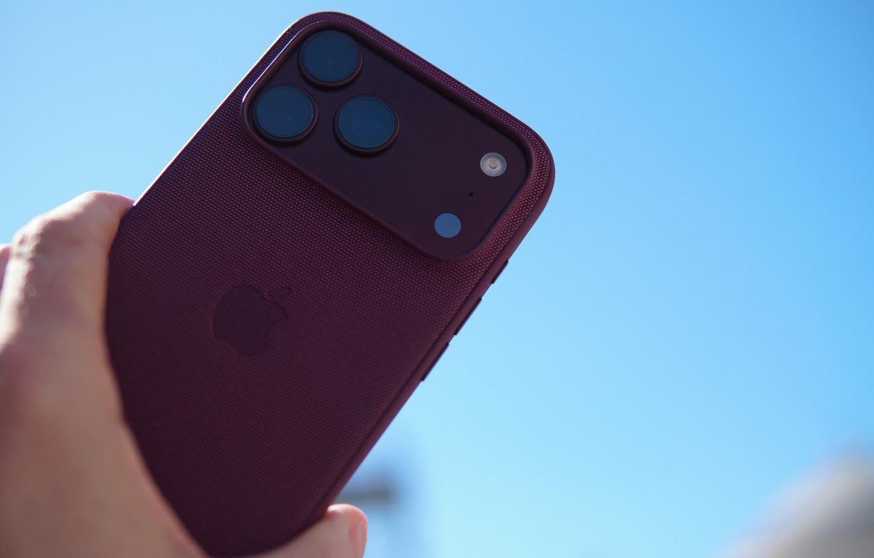 A close-up view of a hand holding a red iPhone with three camera lenses and a flash against a clear blue sky background.