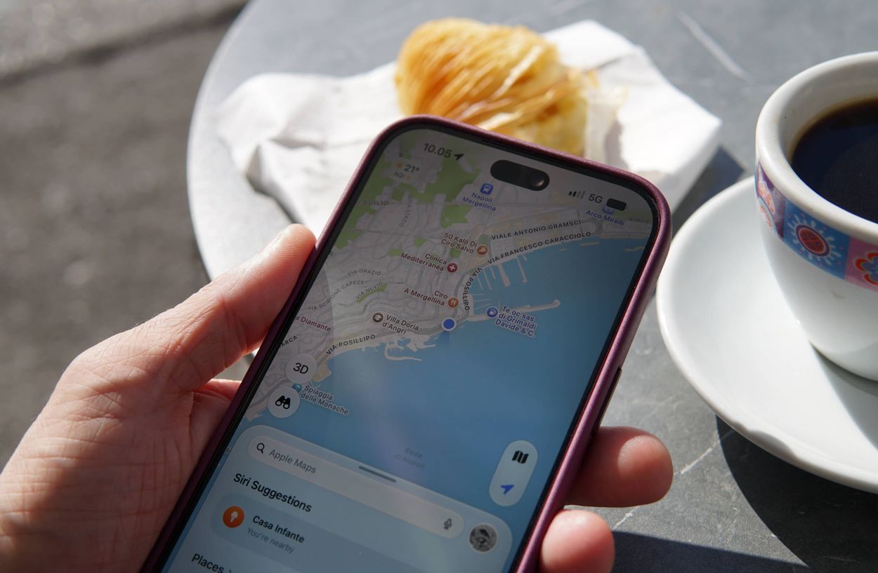 A hand holding a smartphone displaying a map application of a coastal area, with a plate of pastry and a cup of black coffee on a gray table surface nearby.