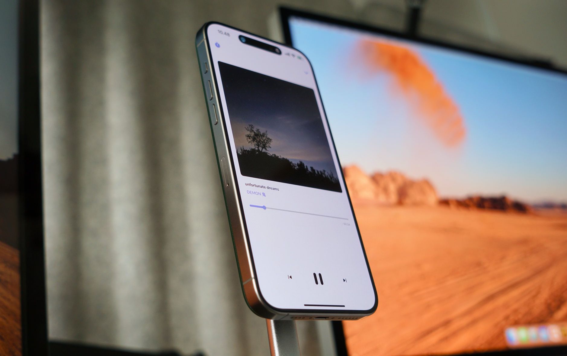 Das Bild zeigt ein iPhone, das Musik abspielt und auf einem Ständer steht. Im Hintergrund ist unscharf ein Wüstenlandschaftsbild auf einem Bildschirm zu sehen.