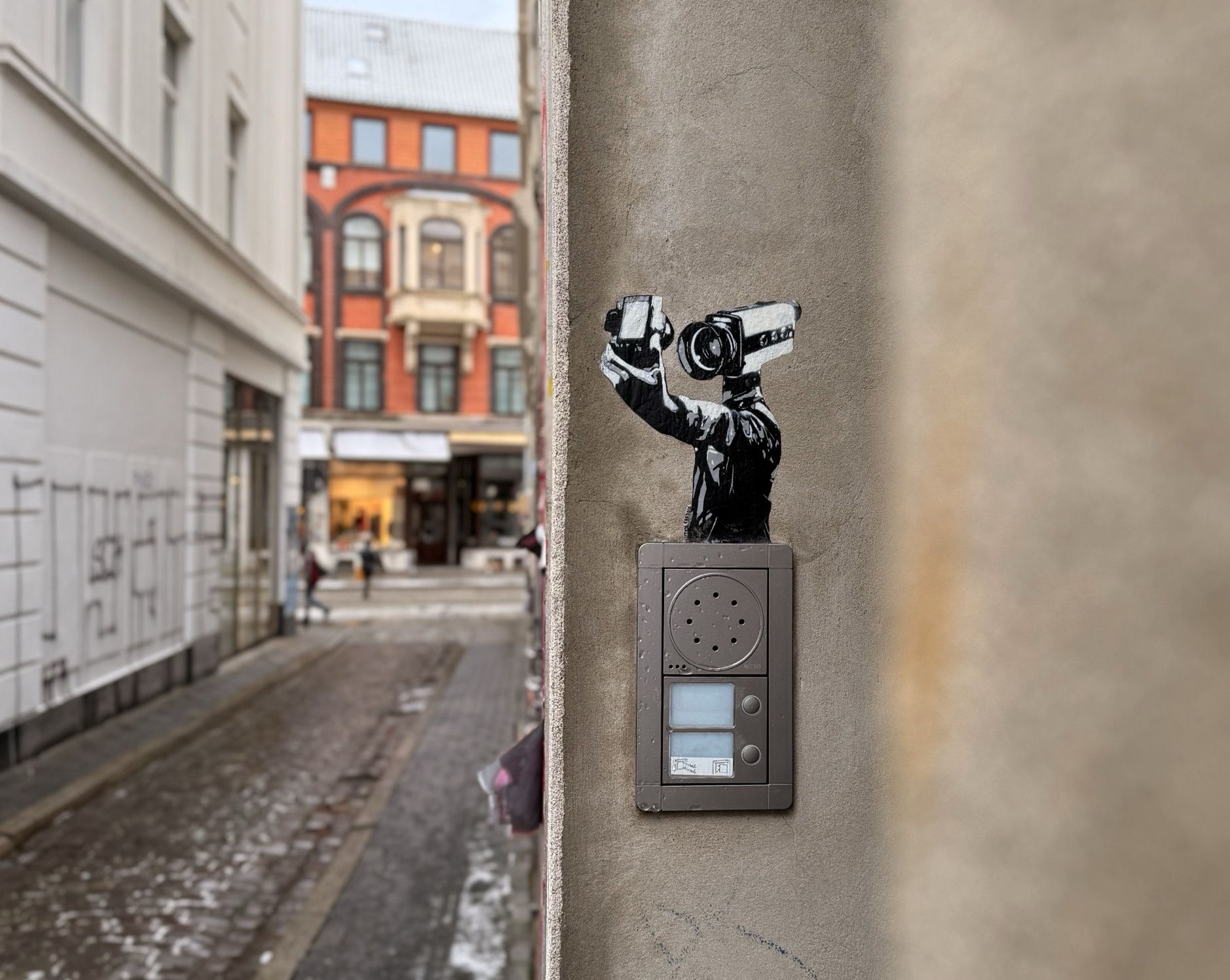 Ein kreatives Stück Street Art zeigt eine schwarz-weiße Figur mit einer Videokamera, die geschickt über einer Gegensprechanlage an einer grauen Hauswand platziert wurde. Im Hintergrund ist eine typisch städtische Straßenszene zu sehen, mit einem auffälligen orangefarbenen Gebäude mit Erkerfenstern. Die künstlerische Installation spielt clever mit der Überwachungsthematik und verwandelt ein alltägliches Objekt in ein nachdenkliches Kunstwerk.