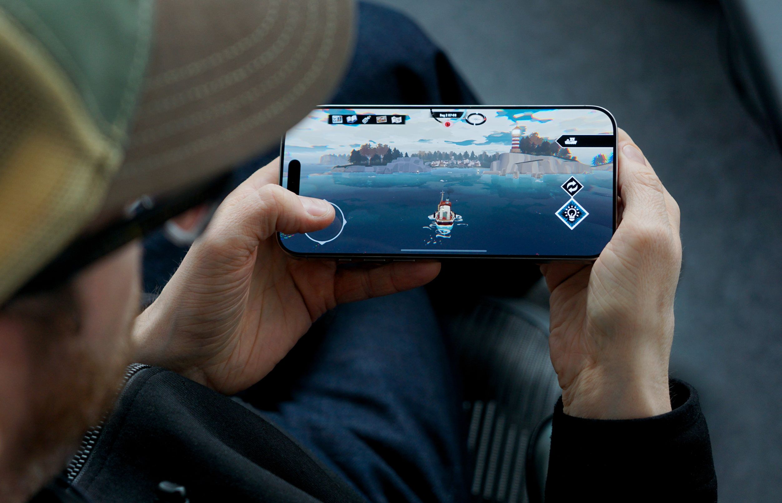 Das Bild zeigt eine Person, die ein Videospiel auf einem iPhone spielt. Auf dem Bildschirm ist ein Boot auf dem Wasser zu sehen, mit einer Küstenlinie und einem Leuchtturm im Hintergrund. Die Person hält das Telefon horizontal mit beiden Händen.