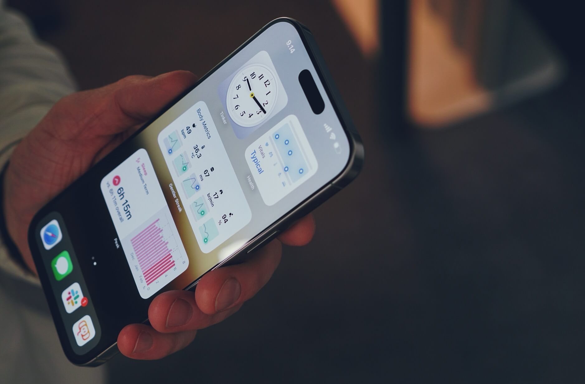 Das Bild zeigt ein iPhone, das in einer Hand gehalten wird. Auf dem Bildschirm sind verschiedene Widgets zu sehen, darunter eine Uhr, Informationen zu Körpermetriken und Schlafdaten. Die Uhrzeit auf dem iPhone ist 9:14 Uhr.