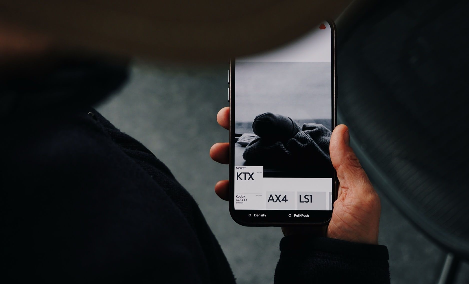 Auf dem Bild hält eine Person ein Smartphone in der Hand, auf dessen Bildschirm eine minimalistische Benutzeroberfläche zu sehen ist. Die Anzeige zeigt ein Foto von einem zusammengerollten Kleidungsstück sowie Optionen wie "KTX", "AX4" und "LS1". Der Stil wirkt modern und schlicht, mit Fokus auf Fotografie und Design.