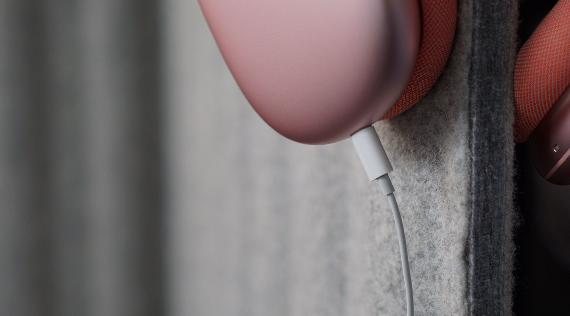 Auf dem Bild ist ein rosa AirPods Max-Kopfhörer zu sehen, der über ein USB-C-Kabel geladen wird. Der Kopfhörer hängt an einer grauen Filzoberfläche. Der Fokus liegt auf dem Ladeanschluss und dem Kabel.
