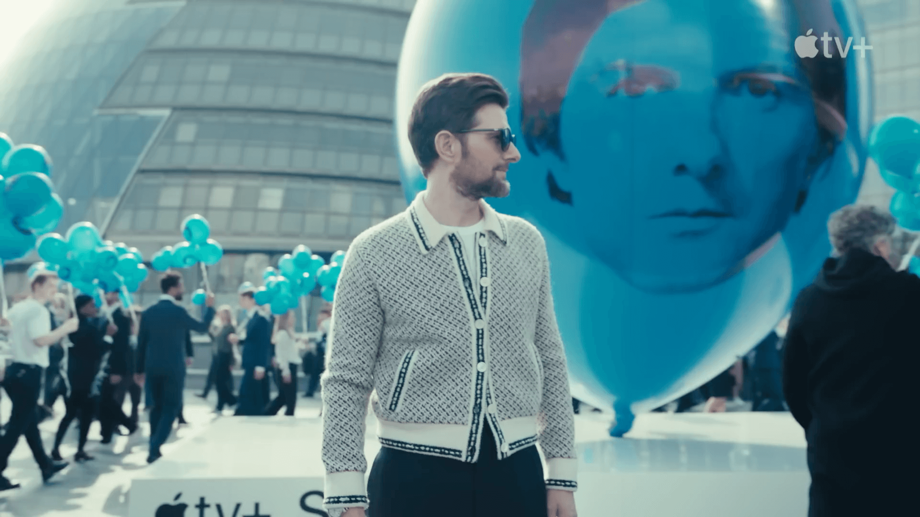 Das Bild zeigt eine Szene aus einer Installation in London, die mit der Apple TV+ Serie Severance verbunden ist. Im Vordergrund steht ein Mann in einem eleganten, gemusterten Cardigan, während im Hintergrund zahlreiche Menschen mit blauen Luftballons zu sehen sind. Die Umgebung ist modern, mit einem Gebäude im Hintergrund und einer riesigen blauen Kugel als Teil der Installation.
