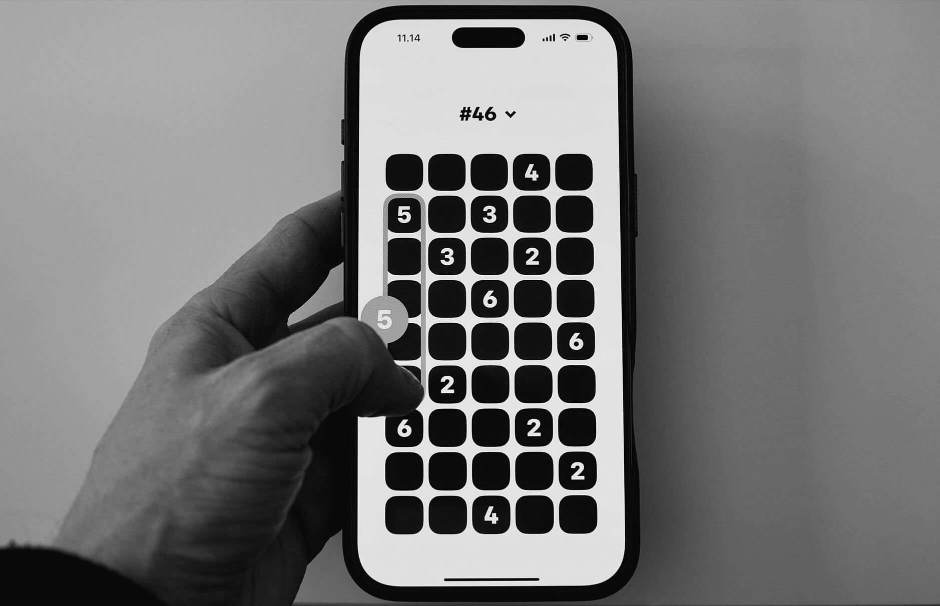 Das Bild zeigt eine Hand, die ein Smartphone hält, auf dem ein minimalistisches Puzzle-Spiel mit Zahlen dargestellt ist. Die schwarzen Felder mit weißen Zahlen scheinen Teil eines logikbasierten Spiels zu sein, bei dem Zahlen durch Wischen bewegt werden. Die Benutzeroberfläche ist schlicht und modern gestaltet.