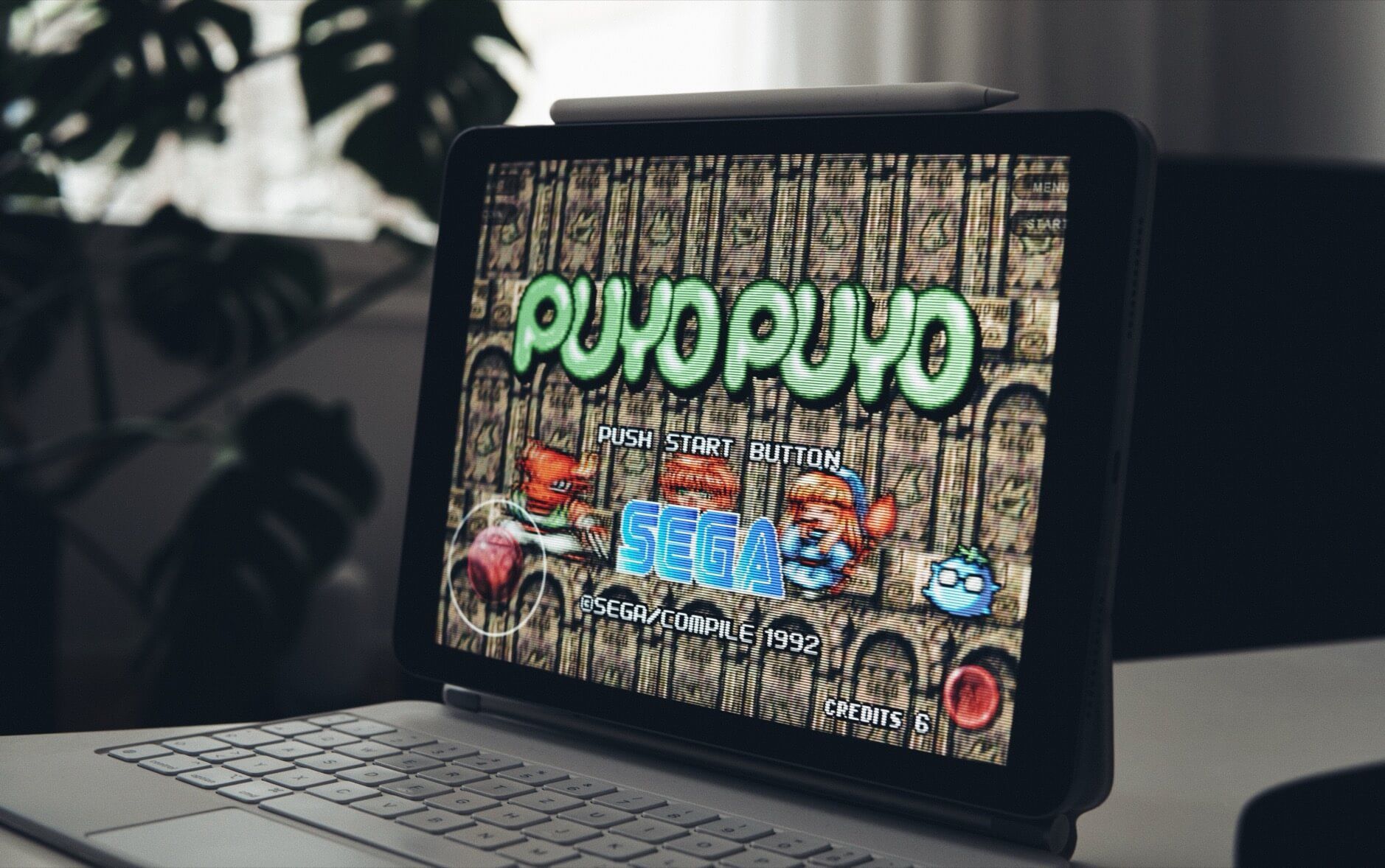Das Bild zeigt ein Tablet mit angeschlossener Tastatur und einem Stift, der oben angebracht ist. Auf dem Bildschirm ist der Startbildschirm des klassischen Videospiels "Puyo Puyo" von SEGA aus dem Jahr 1992 zu sehen. Im Hintergrund ist unscharf eine Pflanze auf einem Schreibtisch erkennbar.