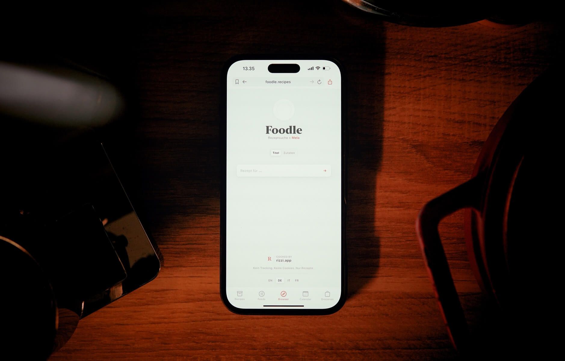 Auf dem Bild sieht man ein Smartphone, das auf einem Holztisch liegt. Auf dem Bildschirm ist die Webseite "Foodle" geöffnet, eine Plattform zur Rezeptsuche. Die Umgebung ist dunkel und das Handy wird von oben beleuchtet.