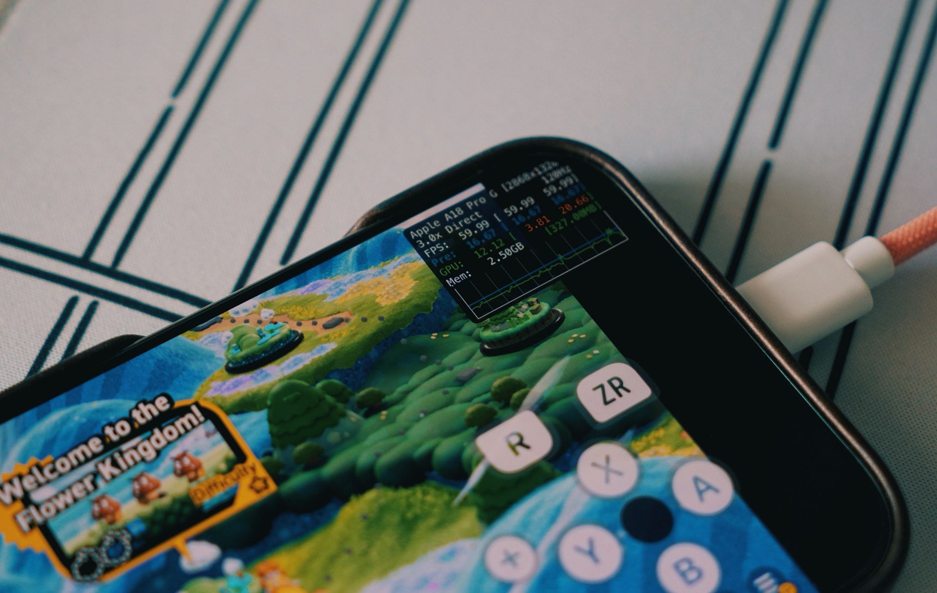 Das Bild zeigt ein Smartphone, auf dem das Spiel "Welcome to the Flower Kingdom!" läuft. Gleichzeitig ist das Gerät über ein Kabel angeschlossen, vermutlich zum Aufladen. Oben rechts auf dem Bildschirm werden technische Leistungsdaten wie FPS und Speichernutzung eingeblendet.