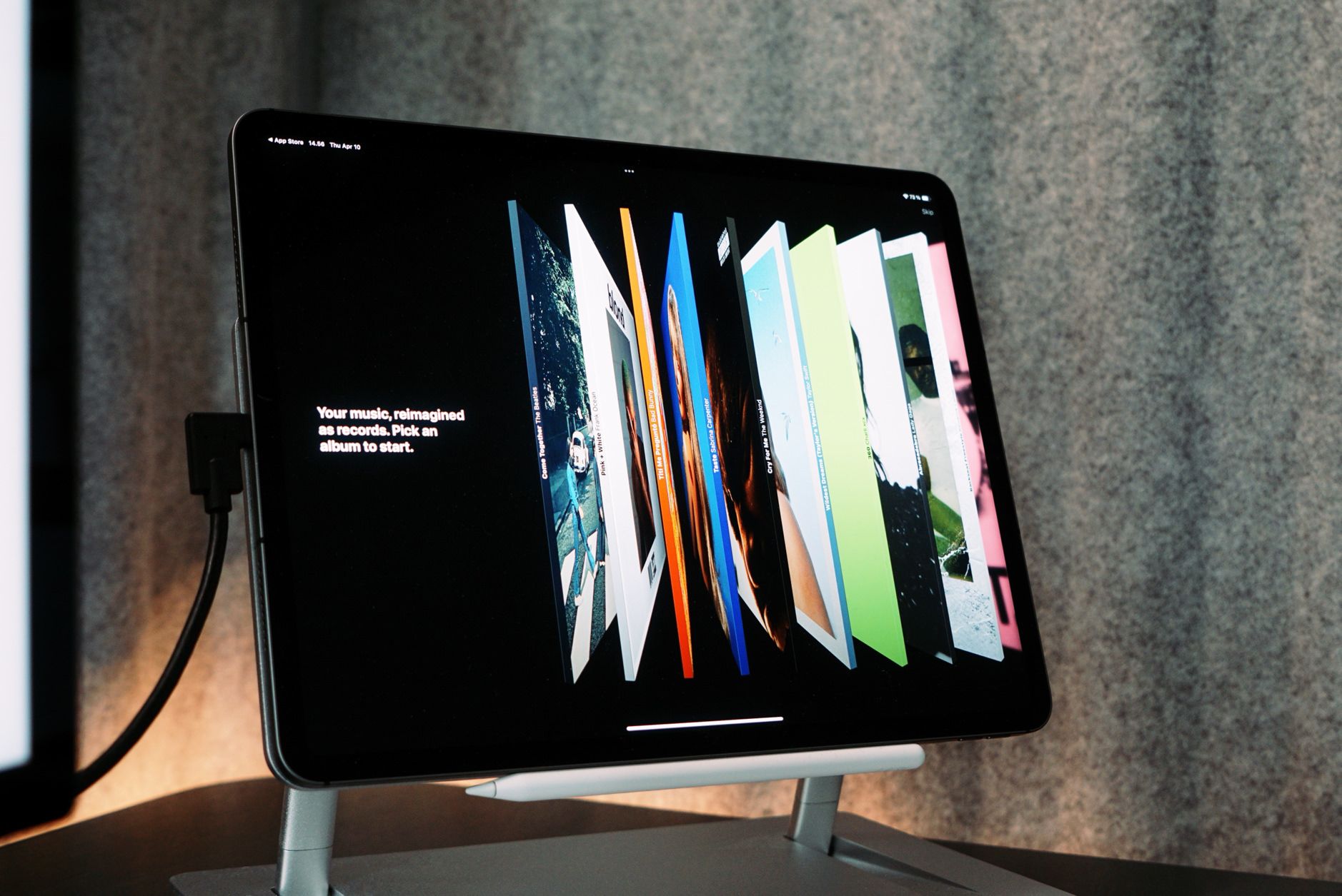 Dieses Bild zeigt ein iPad Pro auf einem Ständer, mit einem Apple Pencil, der magnetisch darunter befestigt ist. Auf dem Bildschirm ist eine Musik-App geöffnet, die verschiedene Albumcover in einer visuellen Darstellung präsentiert, die an Schallplatten erinnert. Ein Text auf dem Display fordert dazu auf, ein Album auszuwählen, um die Musikwiedergabe zu starten.
