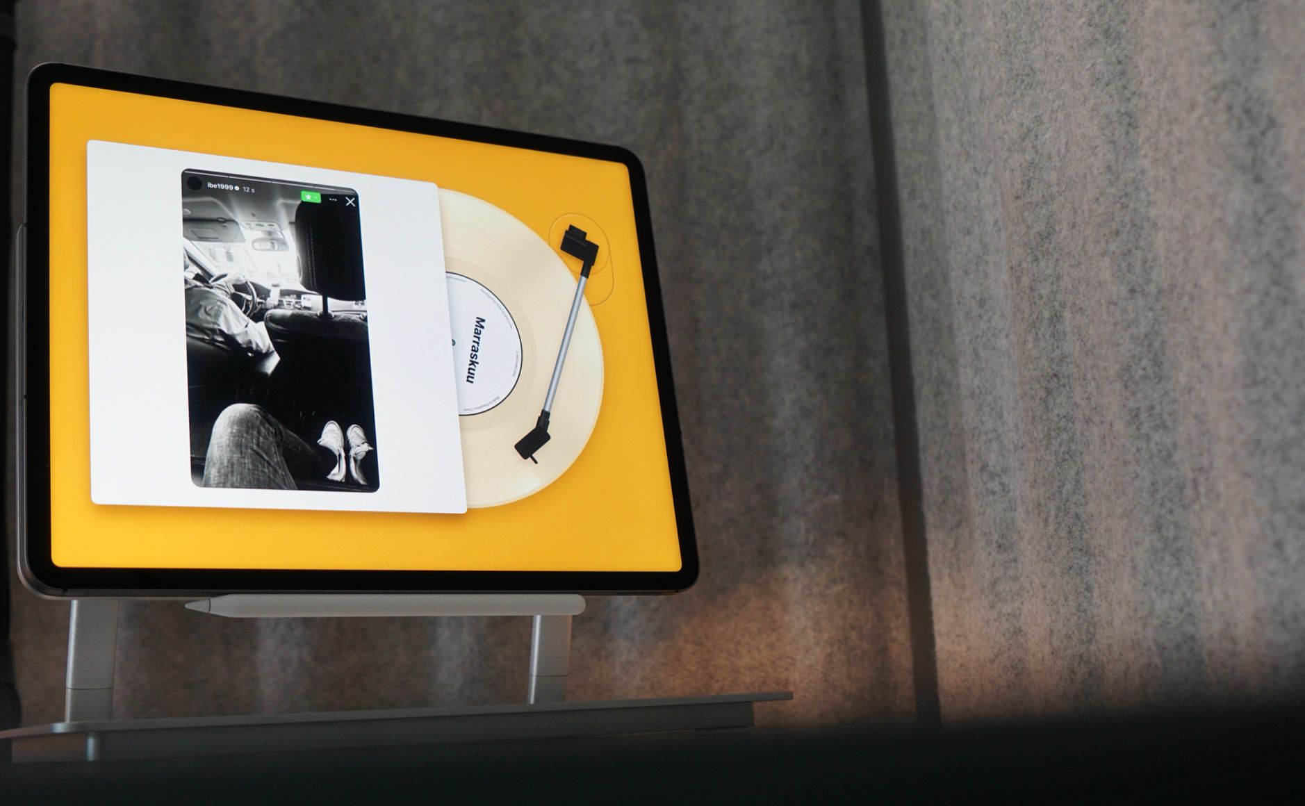 Auf dem Bild ist ein Tablet zu sehen, das auf einem Ständer steht. Der Bildschirm zeigt eine Musikwiedergabe-App, die das Aussehen eines Plattenspielers mit einer Schallplatte und einem Cover nachahmt. Auf der virtuellen Schallplatte steht das Wort "Marraskuu".