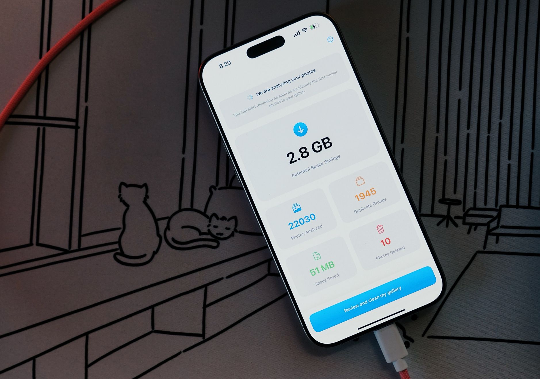 Das Bild zeigt ein Smartphone mit einer Foto-Optimierungs-App, die eine Analyse von 22.030 Fotos durchgeführt hat und ein Potenzial von 2,8 GB Speicherplatzersparnis anzeigt. Die App hat 1945 doppelte Fotogruppen identifiziert, 10 Fotos wurden bereits gelöscht und 51 MB Speicherplatz wurden eingespart. Im Hintergrund ist eine stilisierte Zeichnung von Katzen zu sehen, die auf einer Fensterbank sitzen.