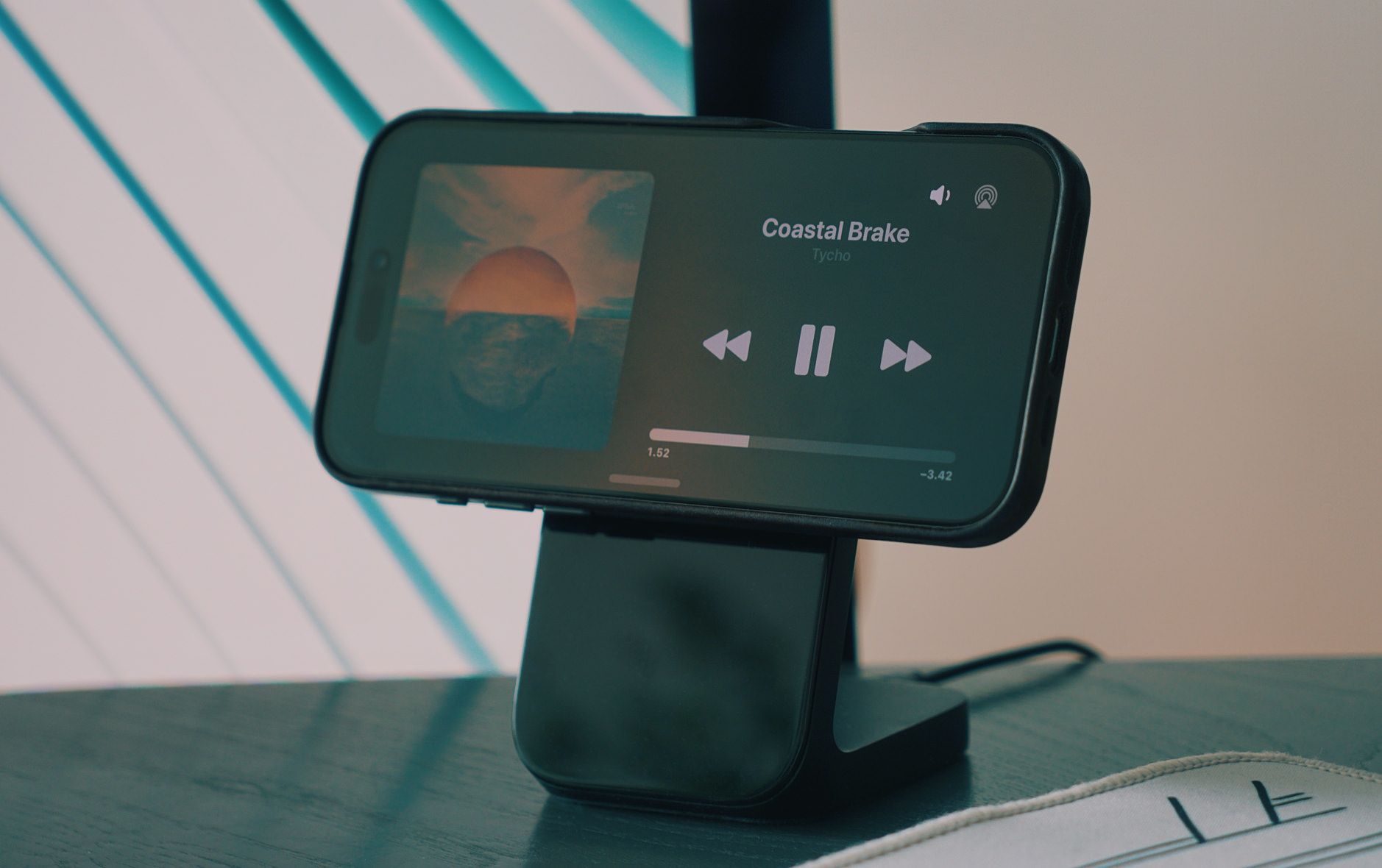 Das Bild zeigt ein Smartphone auf einem speziellen Ständer, das gerade Musik abspielt - den Song "Coastal Brake" von Tycho mit einer Laufzeit von 1:52 Minuten und noch 3:42 Minuten verbleibend. Auf dem Display sind die typischen Mediensteuerungselemente wie Pause, Vor- und Zurück-Tasten sowie ein Fortschrittsbalken zu sehen, während im linken Bereich das Albumcover mit einem Sonnenuntergang über dem Meer angezeigt wird. Das Gerät ist in einer modernen Umgebung platziert, wobei im Hintergrund leichte Streifen zu erkennen sind, die vermutlich von Jalousien oder einer ähnlichen Struktur stammen.
