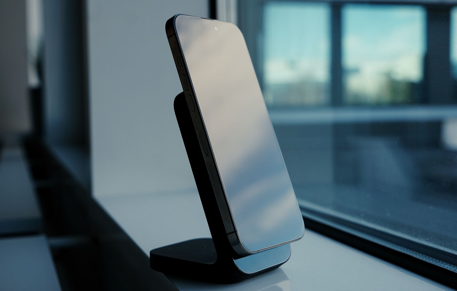 Das Bild zeigt ein modernes Smartphone, das auf einem eleganten, schwarzen Ständer steht. Es befindet sich auf einer Fensterbank in einer Büro- oder Innenraumumgebung, wobei sich die Reflexion des Himmels auf dem Bildschirm spiegelt. Im Hintergrund sind unscharf Fenster mit Blick nach draußen zu erkennen.