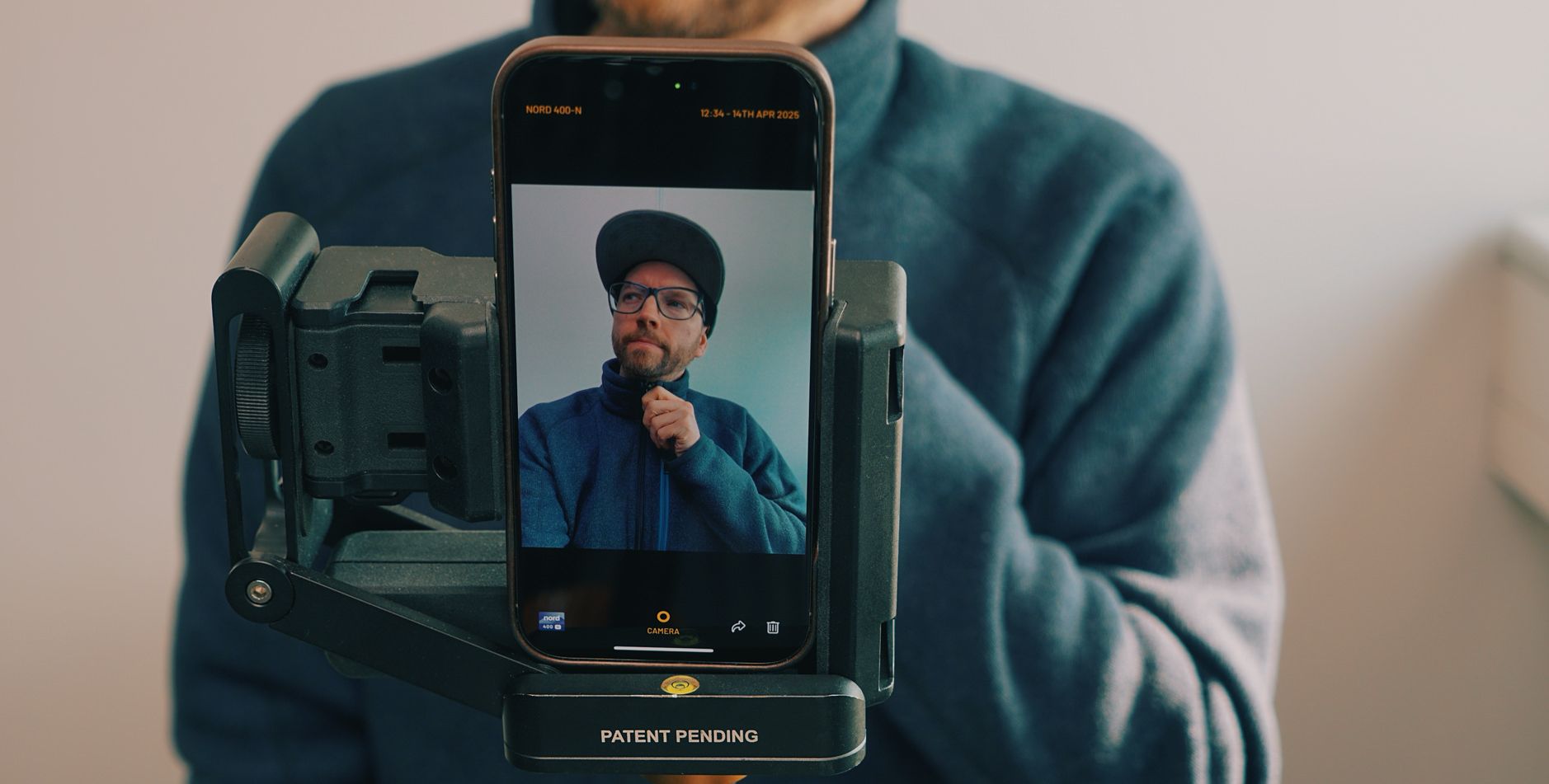 Das Bild zeigt eine Person, die ein Smartphone in einer Halterung oder einem Gimbal hält. Auf dem Bildschirm des Smartphones ist ein Selfie derselben Person zu sehen, die eine Mütze und eine Brille trägt. Die Person im Hintergrund trägt eine blaue Fleecejacke.
