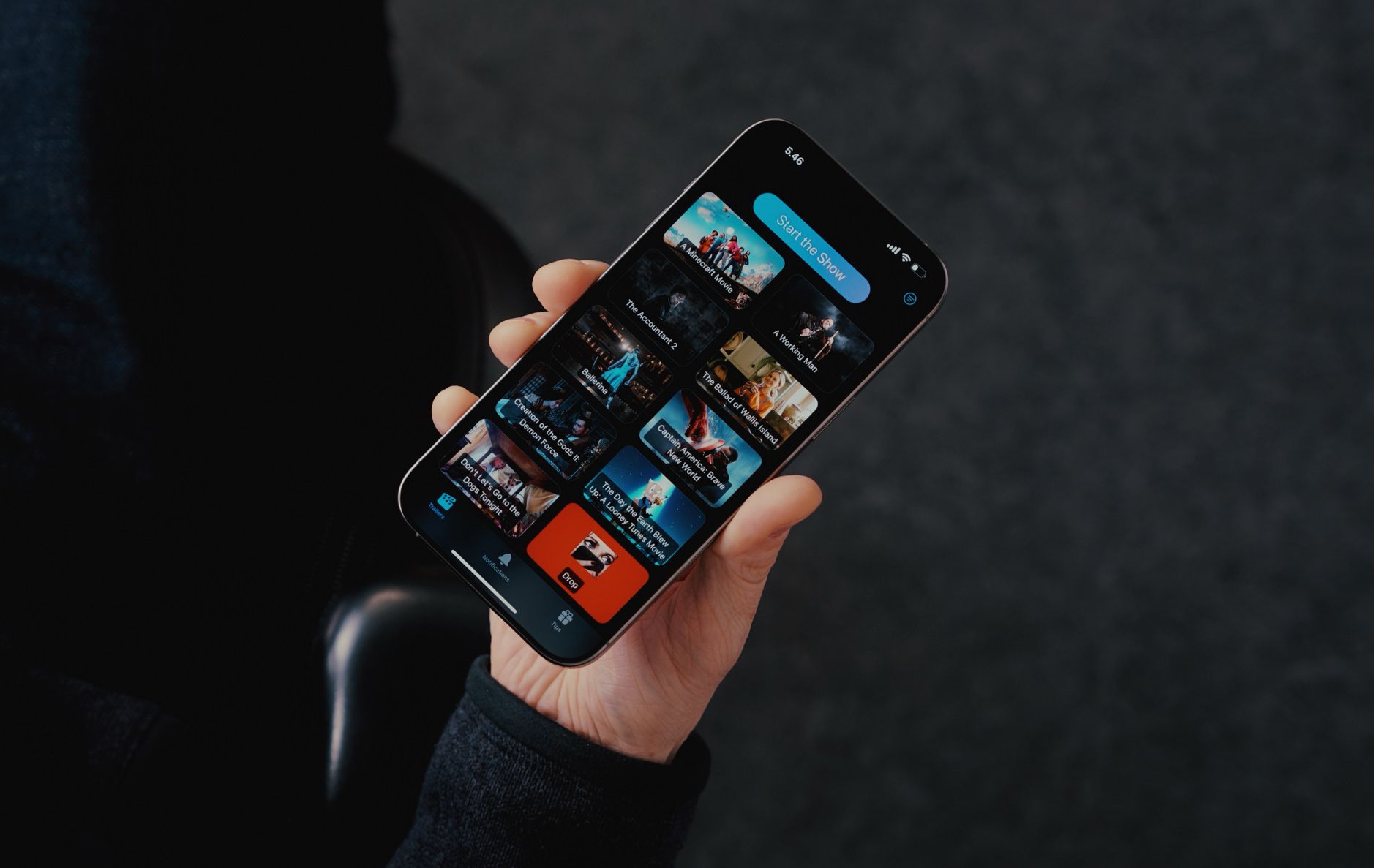 Eine Person hält ein iPhone in der Hand. Auf dem Bildschirm ist die Benutzeroberfläche einer Streaming-App zu sehen, die eine Auswahl verschiedener Filme mit Postern anzeigt. Oben befindet sich ein blauer Button mit der Aufschrift "Start the Show".