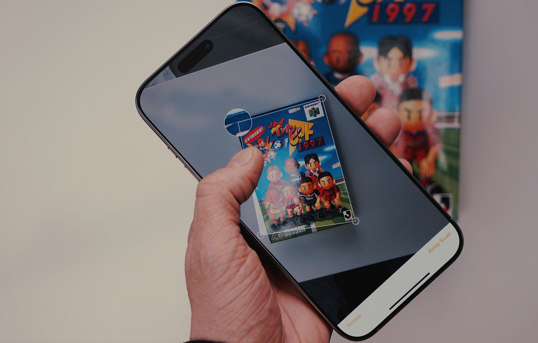 Eine Person hält ein Smartphone in der Hand und scannt damit das Cover eines Videospiels mit japanischer Aufschrift und der Jahreszahl 1997. Auf dem Bildschirm des Smartphones ist das Spielcover deutlich zu erkennen, während das Originalcover im Hintergrund leicht verschwommen sichtbar ist. Unten auf dem Display gibt es die Optionen „Retake“ und „Keep Scan“.