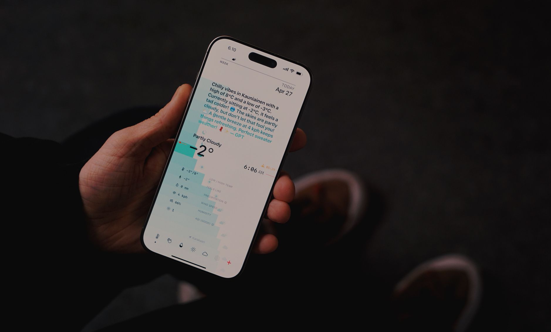 Auf dem Bild ist ein iPhone zu sehen, das die Wetter-App anzeigt. Der Bildschirm zeigt die Wettervorhersage für Kauniainen mit einer Temperatur von -2°C, teilweise bewölktem Himmel und einer poetischen Beschreibung des Wetters am 27. April um 6:06 Uhr morgens. Eine Hand hält das Smartphone vor einem dunklen Hintergrund, wobei die türkisfarbene Benutzeroberfläche der App deutlich zu erkennen ist.