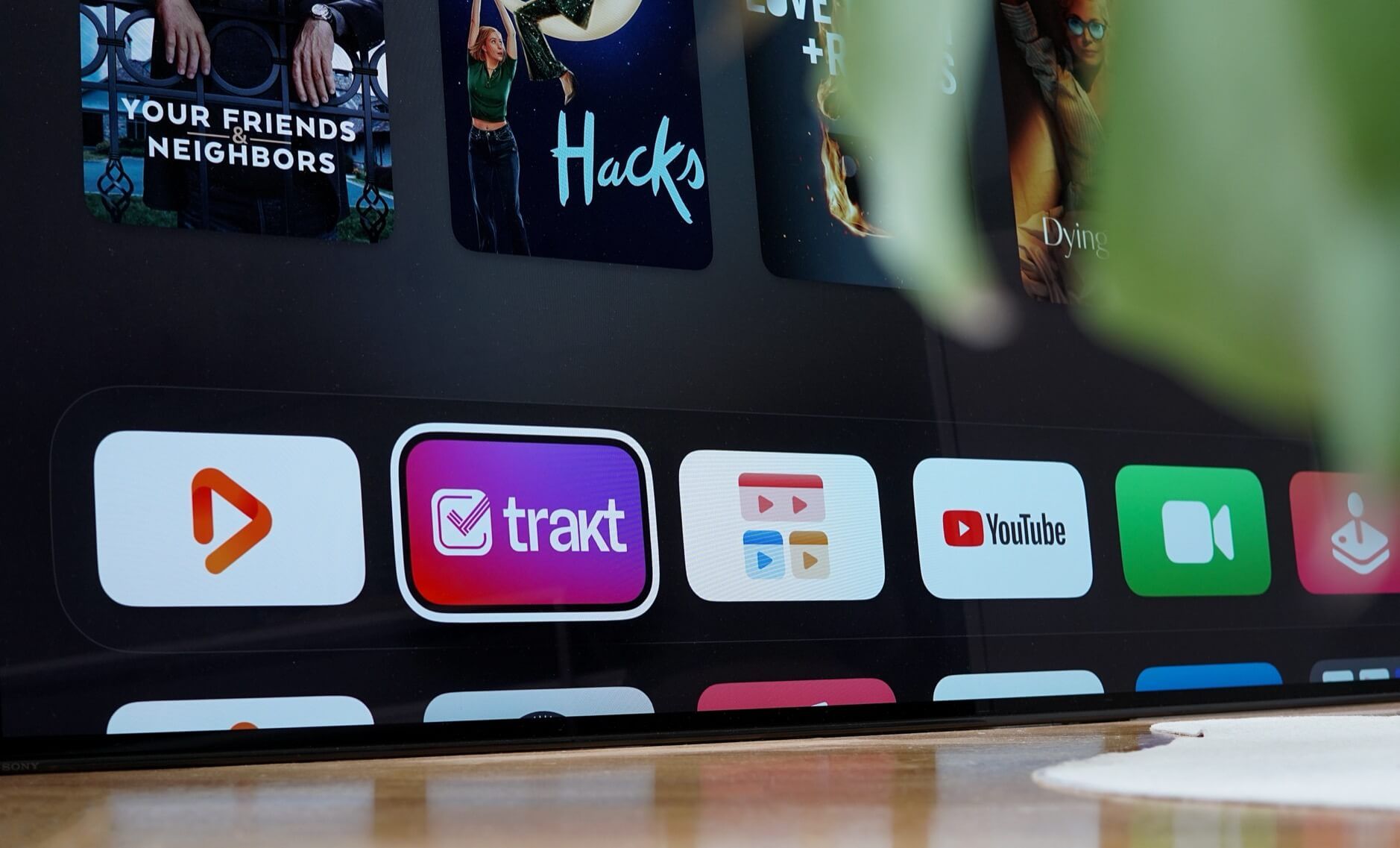 Dieses Bild zeigt einen Apple TV Bildschirm mit verschiedenen App-Icons in der unteren Reihe, darunter Trakt, YouTube und andere Streaming-Dienste. Im oberen Teil des Bildschirms sind Vorschaubilder von Filmen oder Serien zu sehen, wobei eines mit "Hacks" beschriftet ist und ein anderes "YOUR FRIENDS & NEIGHBORS" zeigt. Der Bildschirm scheint auf einem Tisch zu stehen, wobei am unteren Rand des Bildes eine Tischoberfläche zu erkennen ist.