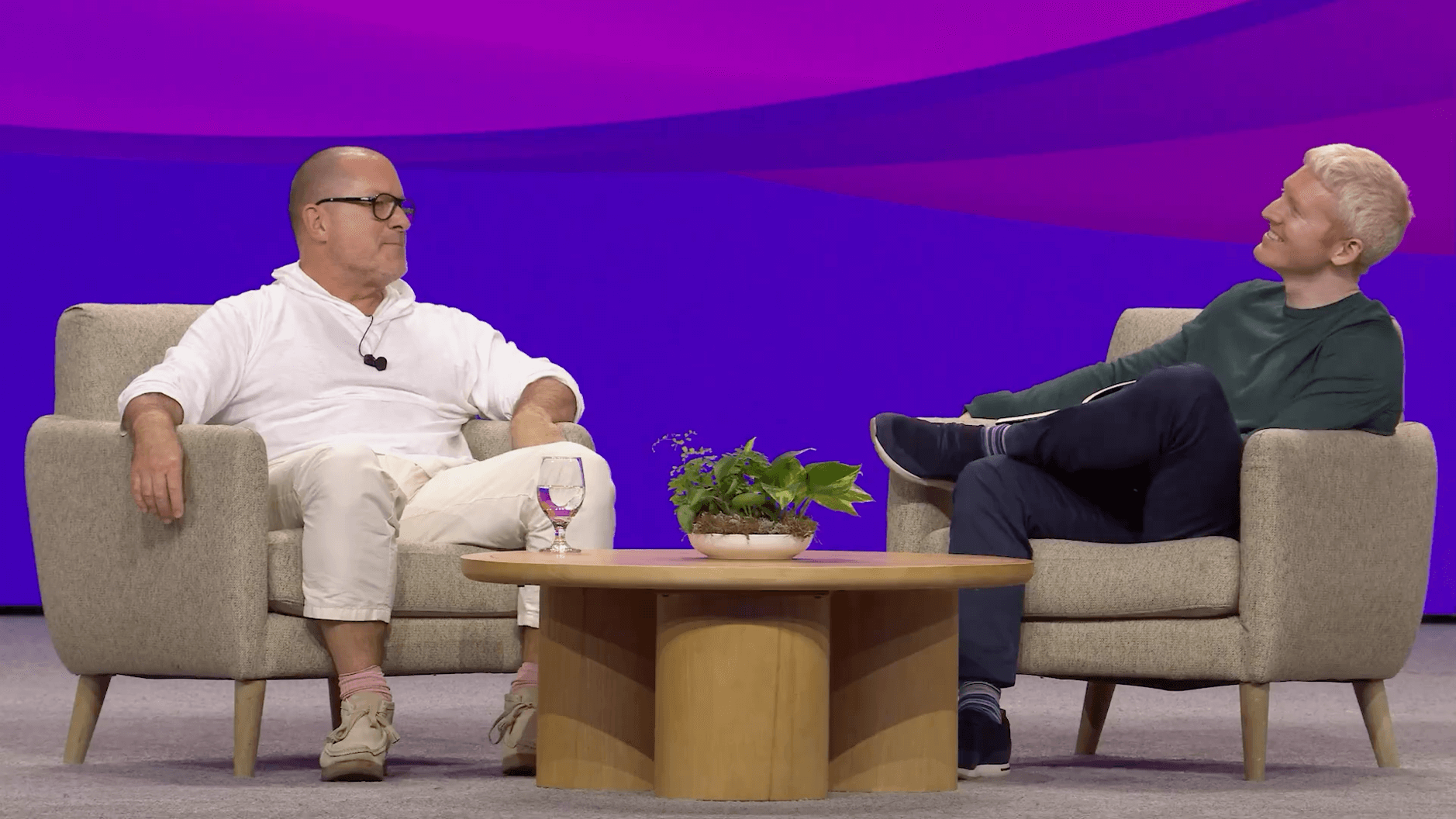 Das Bild zeigt zwei Personen (Jony Ive und Patrick Collison), die in bequemen, beigen Sesseln sitzen und sich in einer Gesprächssituation befinden. Zwischen ihnen steht ein runder Holztisch mit einer kleinen Pflanze und einem Glas, während der Hintergrund in kräftigen Lila- und Violetttönen gestaltet ist. Die Person links trägt ein weißes Outfit mit hellen Schuhen, während die Person rechts in dunkler Kleidung zu sehen ist.