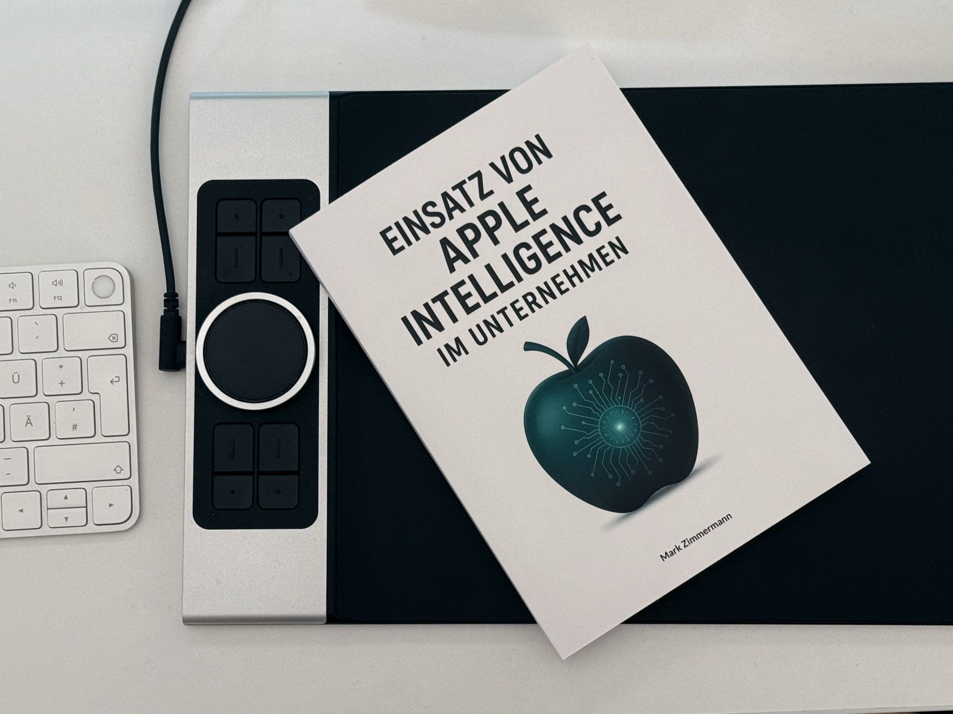 Das Bild zeigt ein Buch mit dem Titel "EINSATZ VON APPLE INTELLIGENCE IM UNTERNEHMEN" von Mark Zimmermann, welches auf einer Arbeitsfläche neben einer Apple-Tastatur und einem Loupedeck-Controller liegt. Auf dem Buchcover ist ein stilisierter Apfel mit digitalen Schaltkreisen zu sehen, der die Verbindung zwischen Apple und künstlicher Intelligenz symbolisiert. Die moderne Arbeitsumgebung mit Apple-Zubehör unterstreicht den thematischen Fokus des Buches auf Apple-Technologie im Unternehmenskontext.