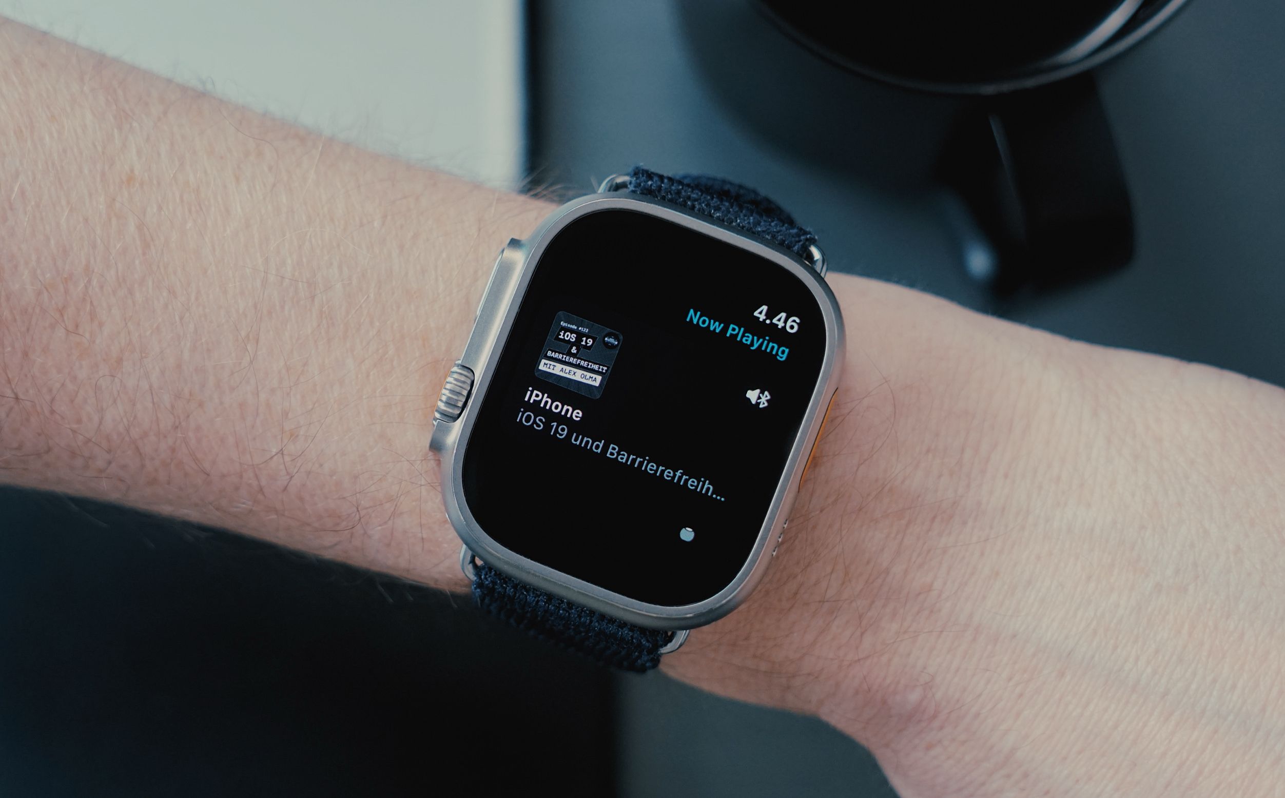 Auf dem Bild ist ein Handgelenk zu sehen, an dem eine Apple Watch getragen wird. Der Bildschirm zeigt aktuelle Wiedergabedetails, einschließlich des Titels und des Podcasts "iOS 19 und Barrierefreiheit". Im Hintergrund sind eine Tasse und eine Arbeitsfläche sichtbar.