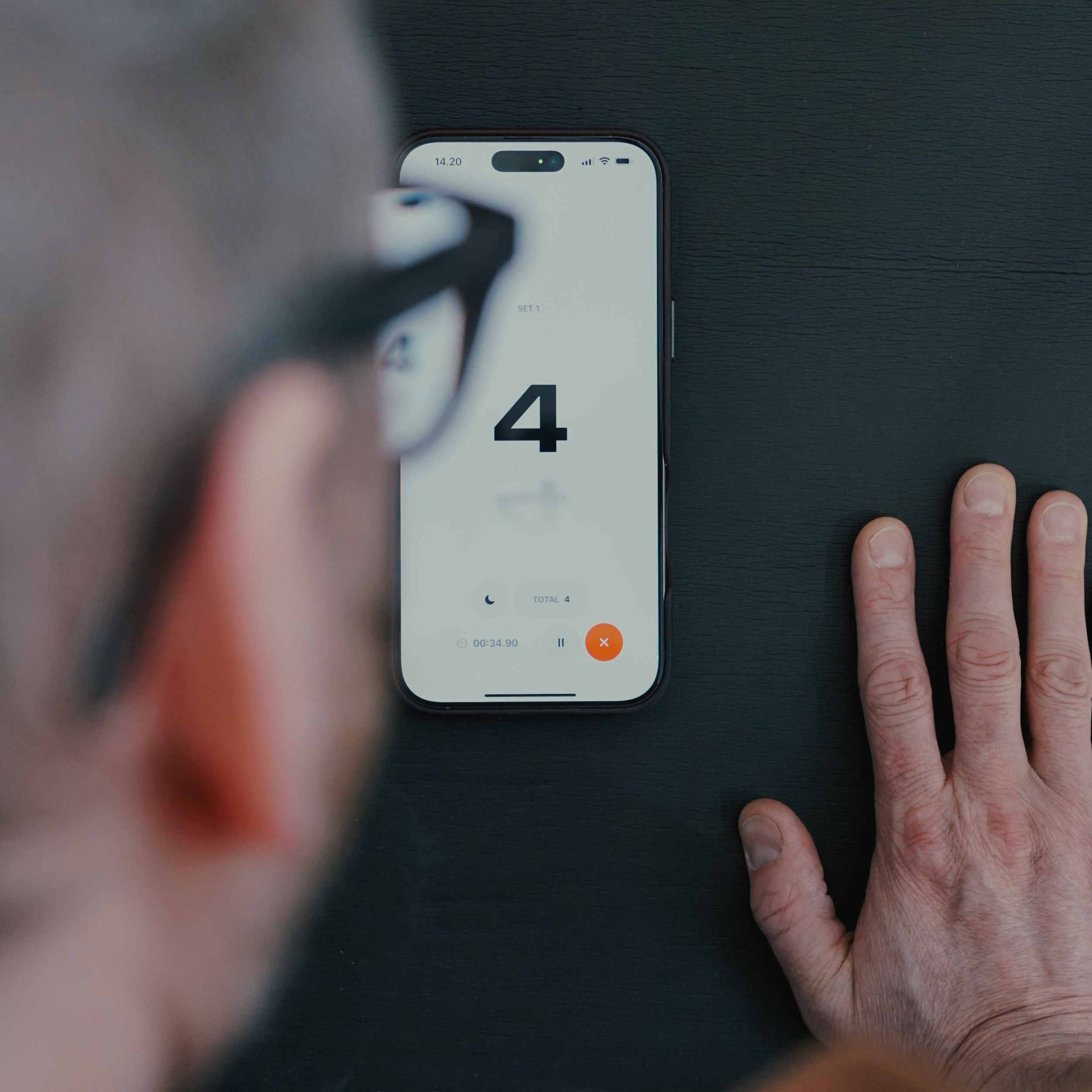 Auf dem Bild sieht man ein Smartphone, das auf einem Yoga-Matte liegt. Der Bildschirm zeigt die Zahl 4 an, während eine Hand daneben auf der Tischoberfläche ruht. Im Hintergrund sieht man die verschwommene Silhouette eines Mannes mit Brille.