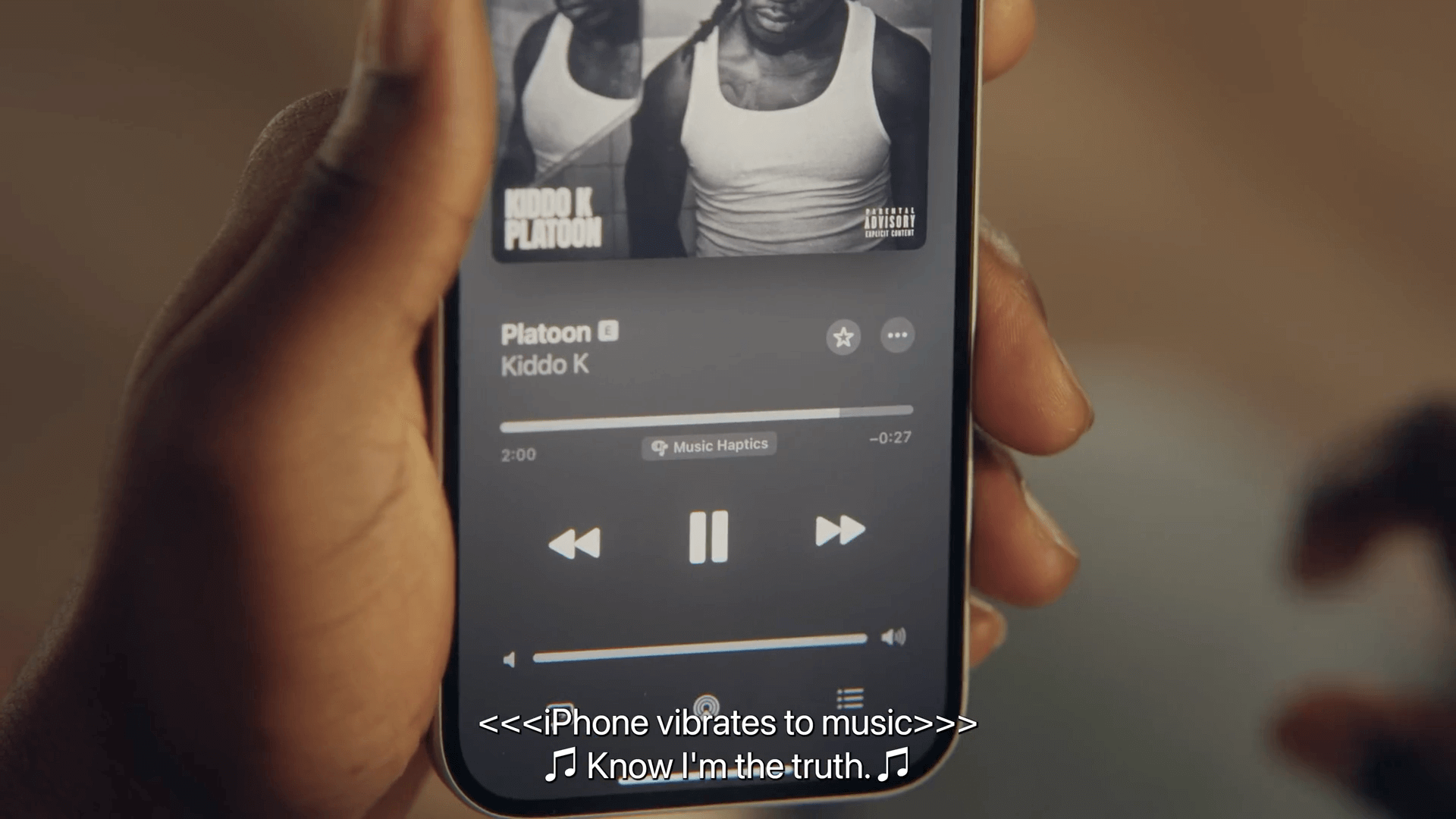Auf dem Bild sieht man eine Hand, die ein iPhone hält, auf dem ein Musikstreaming-Dienst geöffnet ist. Der Bildschirm zeigt das Lied "Platoon" von Kiddo K und die Musik spielt im Hintergrund. Zusätzlich vibriert das iPhone, während der Nutzer den Text "Know I'm the truth" hört.