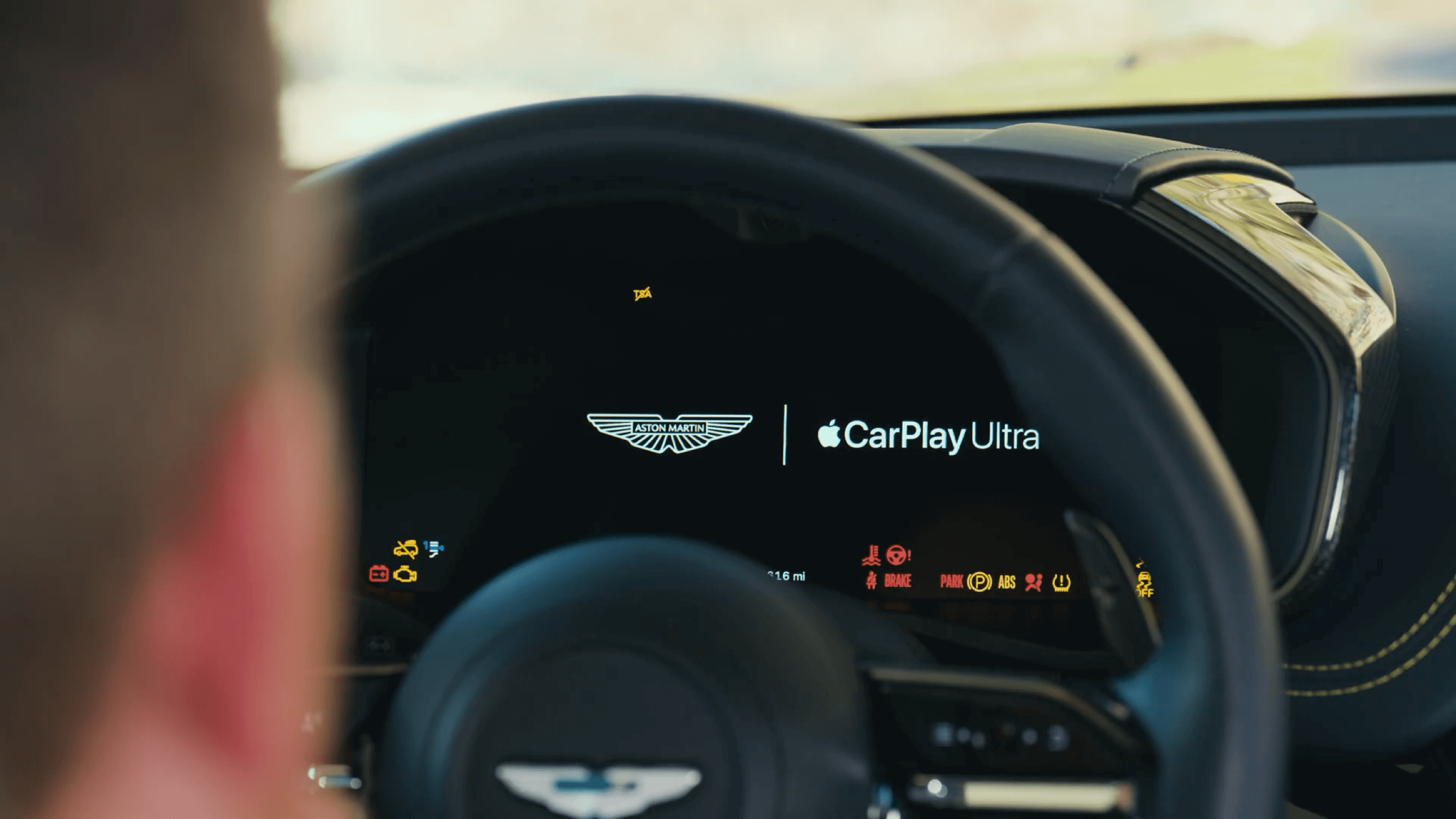 Das Bild zeigt das Armaturenbrett eines Fahrzeugs, auf dem das Logo von Aston Martin und die Anzeige für Apple CarPlay Ultra zu sehen sind. Im Hintergrund erkennt man einige Warnlämpchen und Informationen zum Fahrzeugstatus. Der Fahrer ist im Vordergrund leicht sichtbar, während er auf das Display schaut.