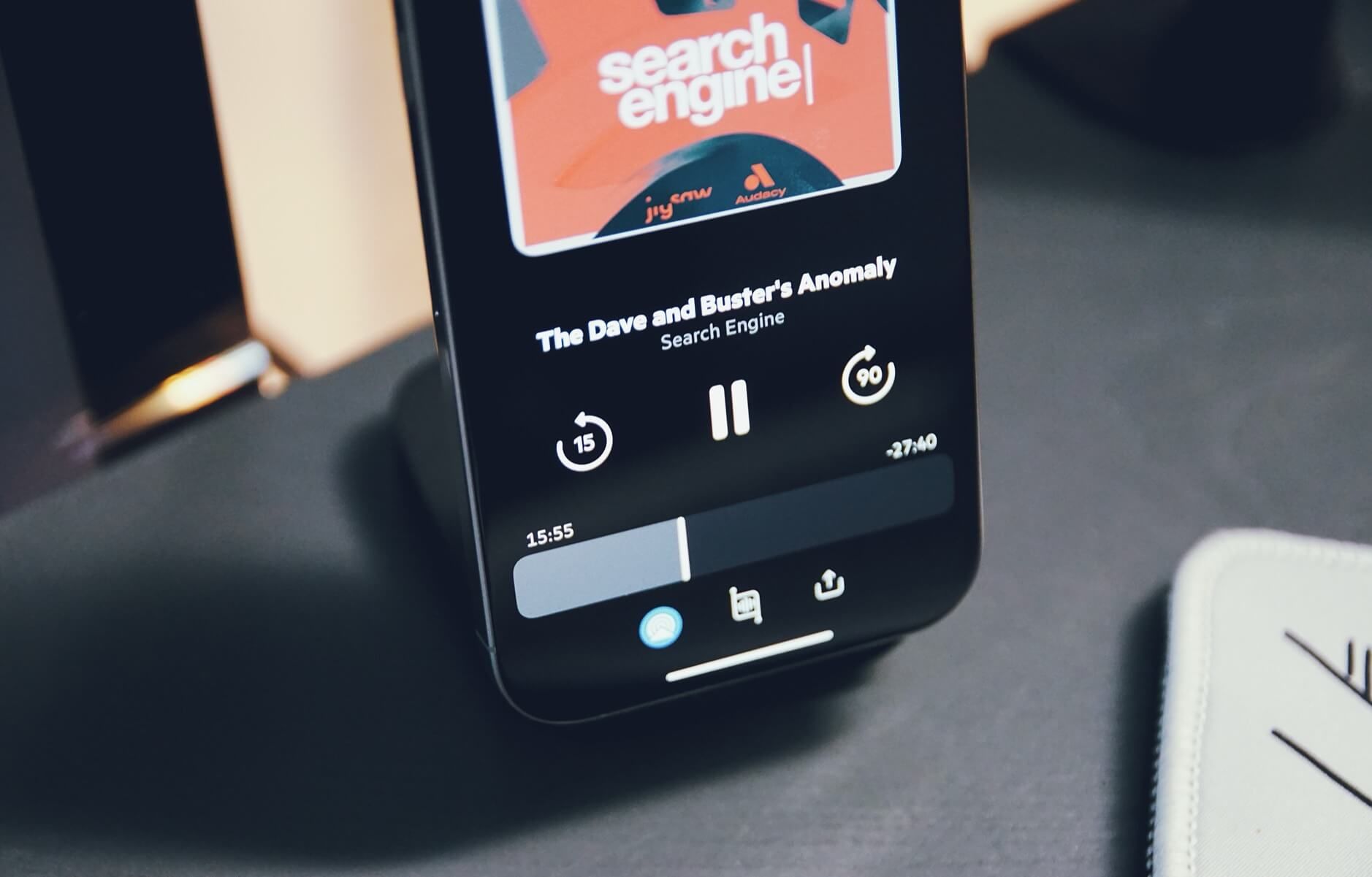 Das Bild zeigt ein Smartphone, auf dem ein Podcast namens "The Dave and Buster's Anomaly Search Engine" abgespielt wird. Der Podcast-Player zeigt die Wiedergabesteuerung mit Pause-Taste, Vor- und Zurückspulen-Optionen sowie einen Fortschrittsbalken, der anzeigt, dass 15:55 Minuten vergangen sind und noch 27:40 Minuten verbleiben. Im oberen Bereich des Bildschirms ist ein orangefarbenes Podcast-Cover mit dem Text "search engine" und den Logos von jnyhw und Audacy zu sehen.