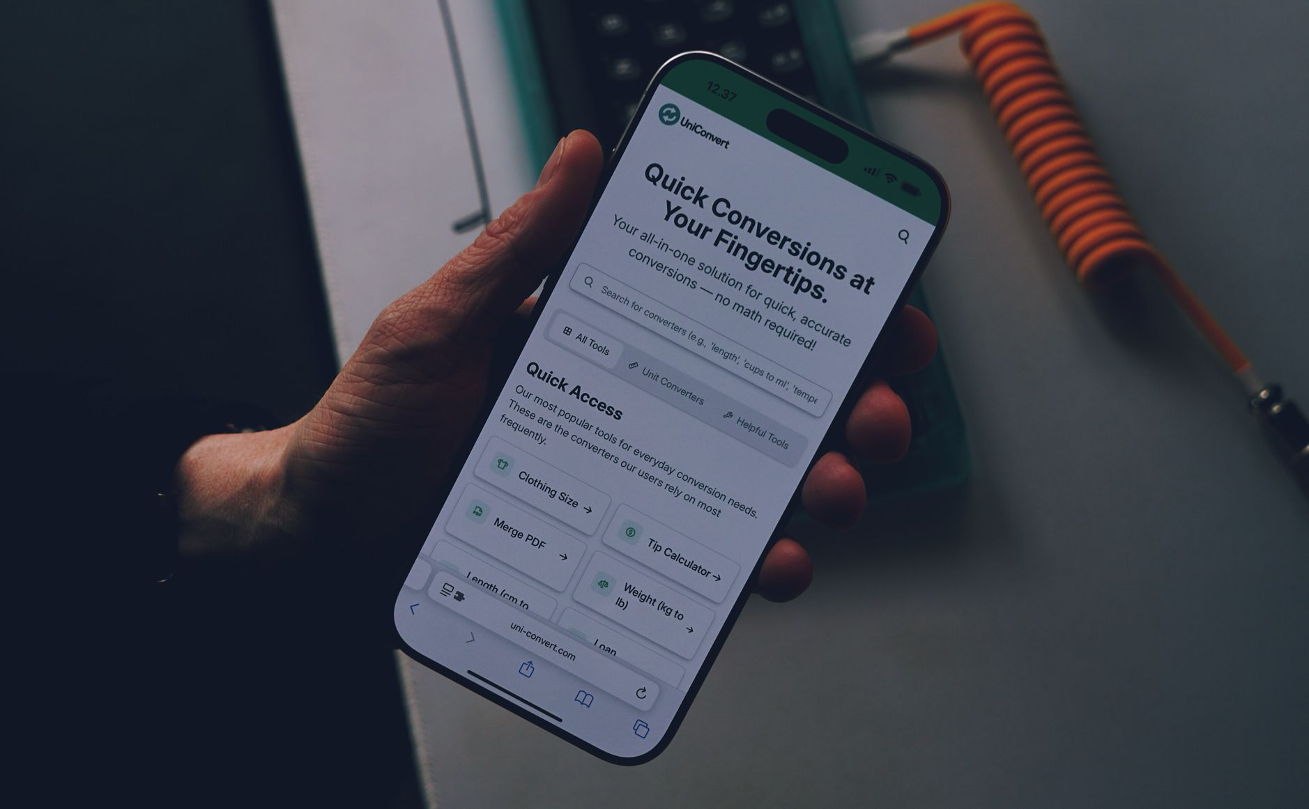 Das Bild zeigt ein Smartphone, auf dem die "UniConvert" App geöffnet ist, die schnelle und genaue Umrechnungen ohne mathematische Kenntnisse anbietet. Auf dem Bildschirm sieht man verschiedene Umrechnungsoptionen wie Kleidergrößen, PDF-Zusammenführung, Längenumrechnung und Gewichtsumrechnung unter der Überschrift "Quick Access". Im Hintergrund sind verschwommen einige technische Geräte zu erkennen, darunter ein orangefarbenes Kabel.