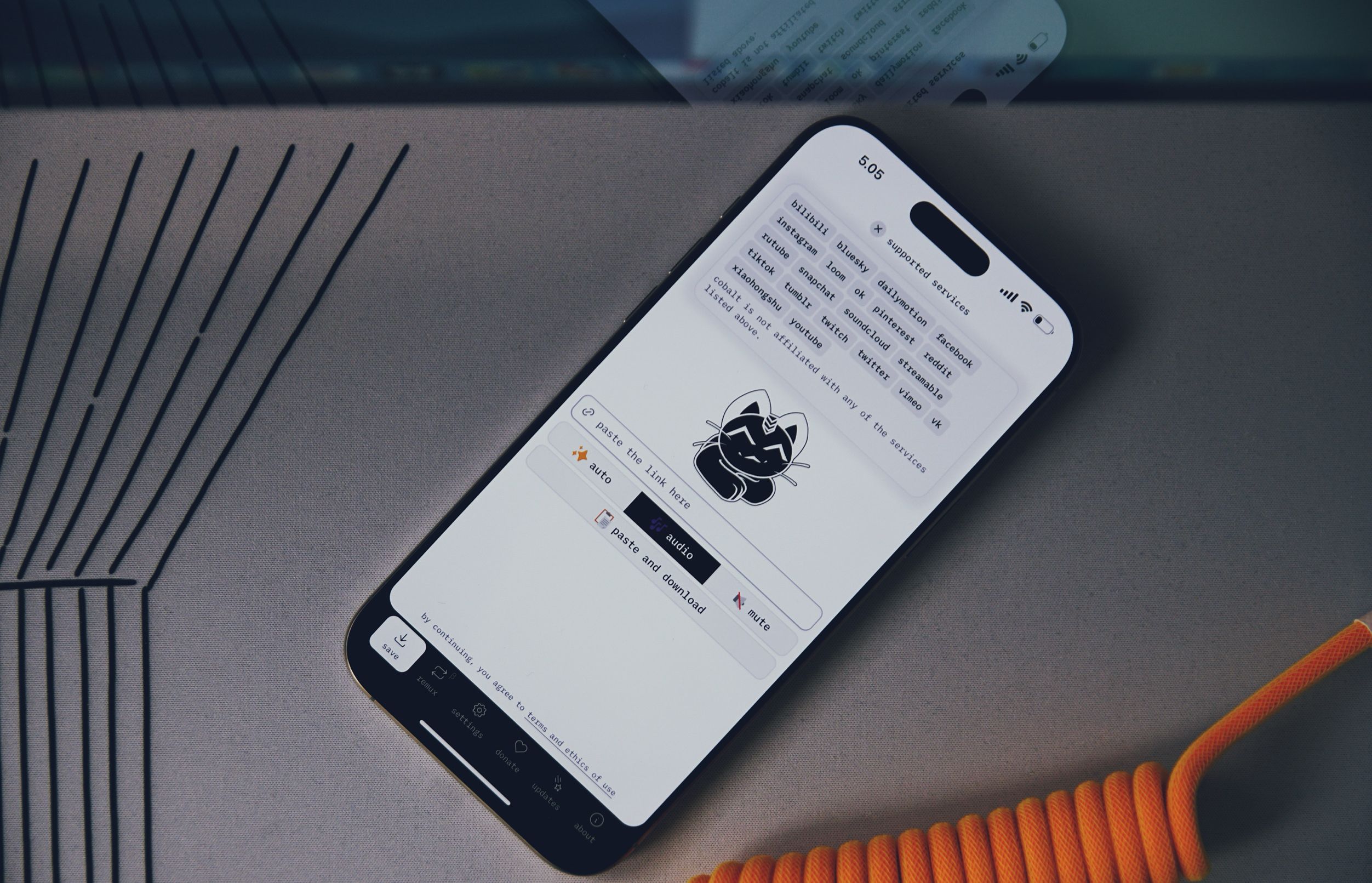 Auf dem Bild liegt ein Smartphone auf einer glatten Oberfläche. Der Bildschirm zeigt eine Benutzeroberfläche, die verschiedene unterstützte Dienste auflistet, darunter Plattformen wie TikTok und YouTube. Neben dem Smartphone ist ein orangefarbener Kabel mit spiralförmiger Struktur zu sehen.
