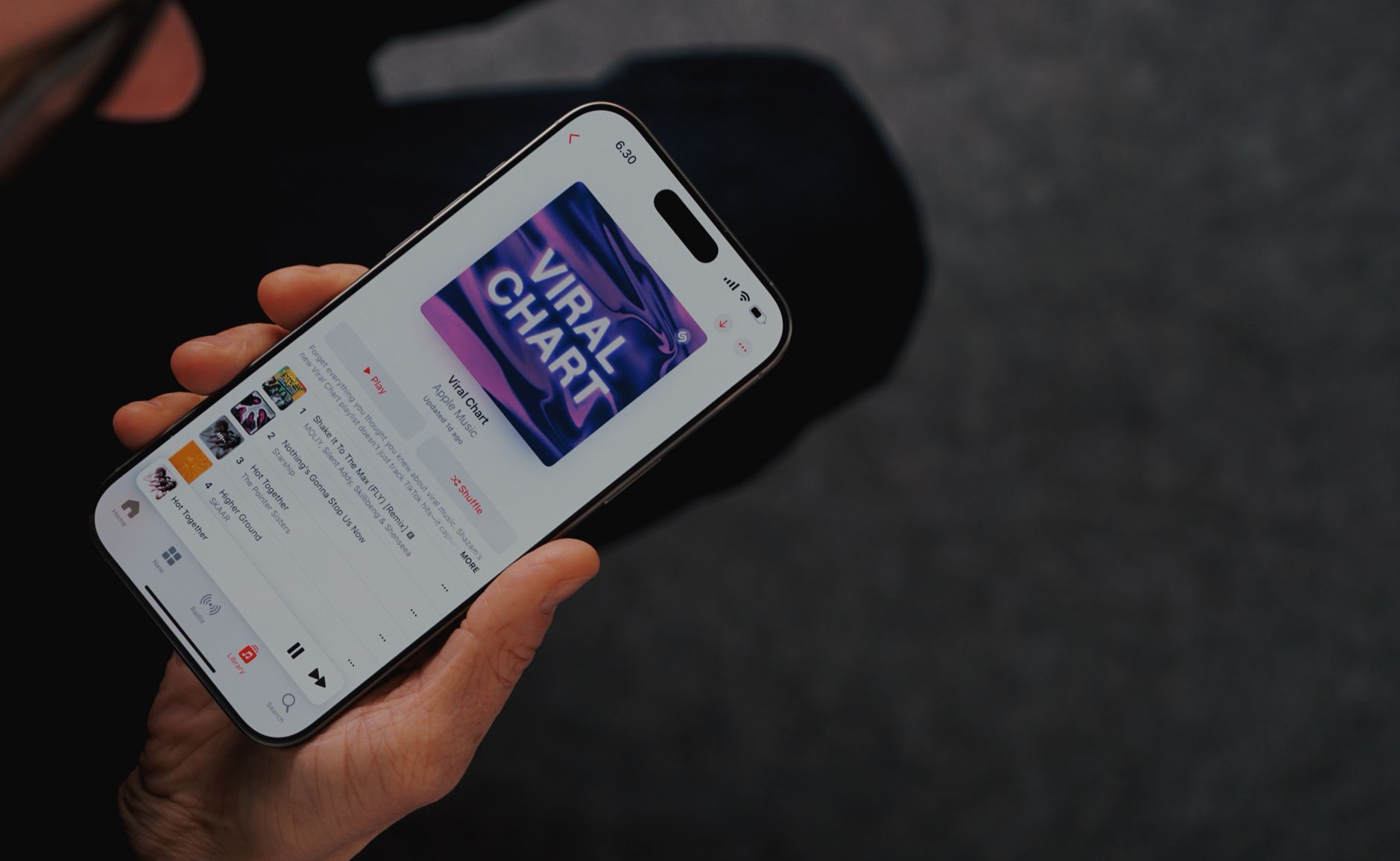 Ein Smartphone zeigt die Musik-App mit dem "Viral Chart" auf dem Display. Die Person hält das Gerät in der Hand und scheint durch eine Playlist oder Musikauswahl zu navigieren. Die Benutzeroberfläche der App ist klar erkennbar mit verschiedenen Musiktiteln in einer Liste unter dem hervorgehobenen "Viral Chart"-Banner.