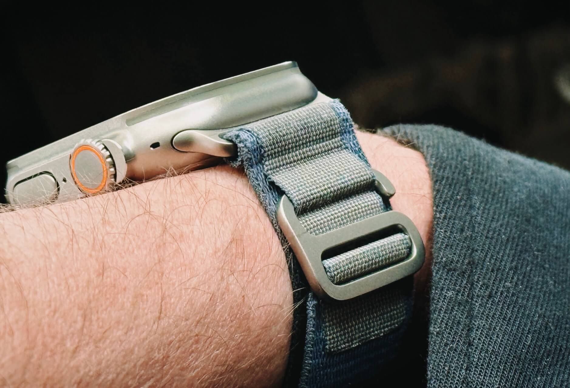 Close-up of a silver smartwatch with an orange-accented rotating crown, secured to a wrist by a gray and blue fabric strap with a brushed metal clasp. The person's arm and dark clothing are also visible.

