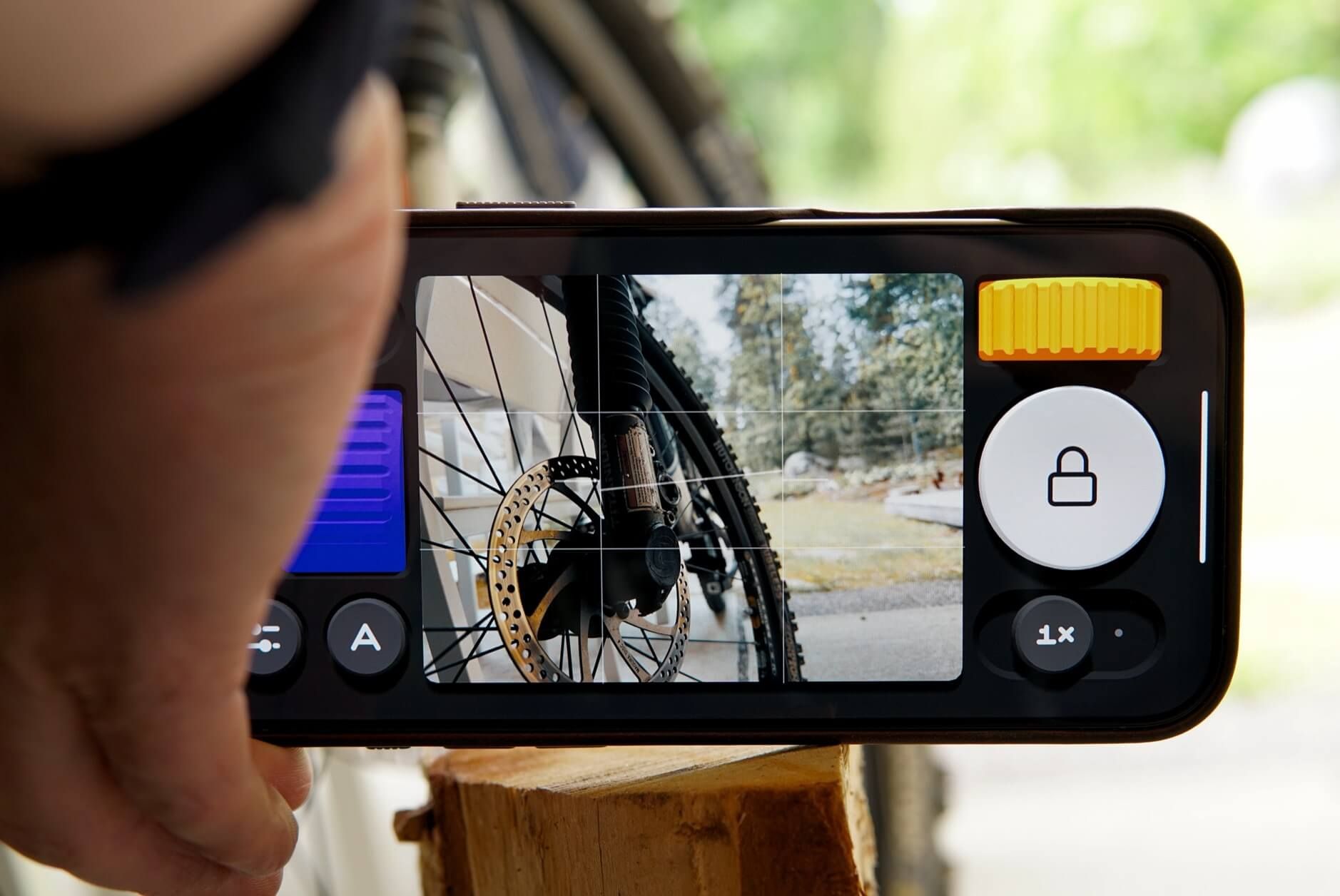 Auf dem Bild hält eine Hand ein Smartphone, das die Kamera-App geöffnet hat. Auf dem Bildschirm ist das Vorderrad eines Fahrrads mit Fokus auf die Speichen und die Bremsscheibe zu sehen. Auffällig sind das gelbe Einstellrad und das große Schlosssymbol auf der rechten Seite der App.