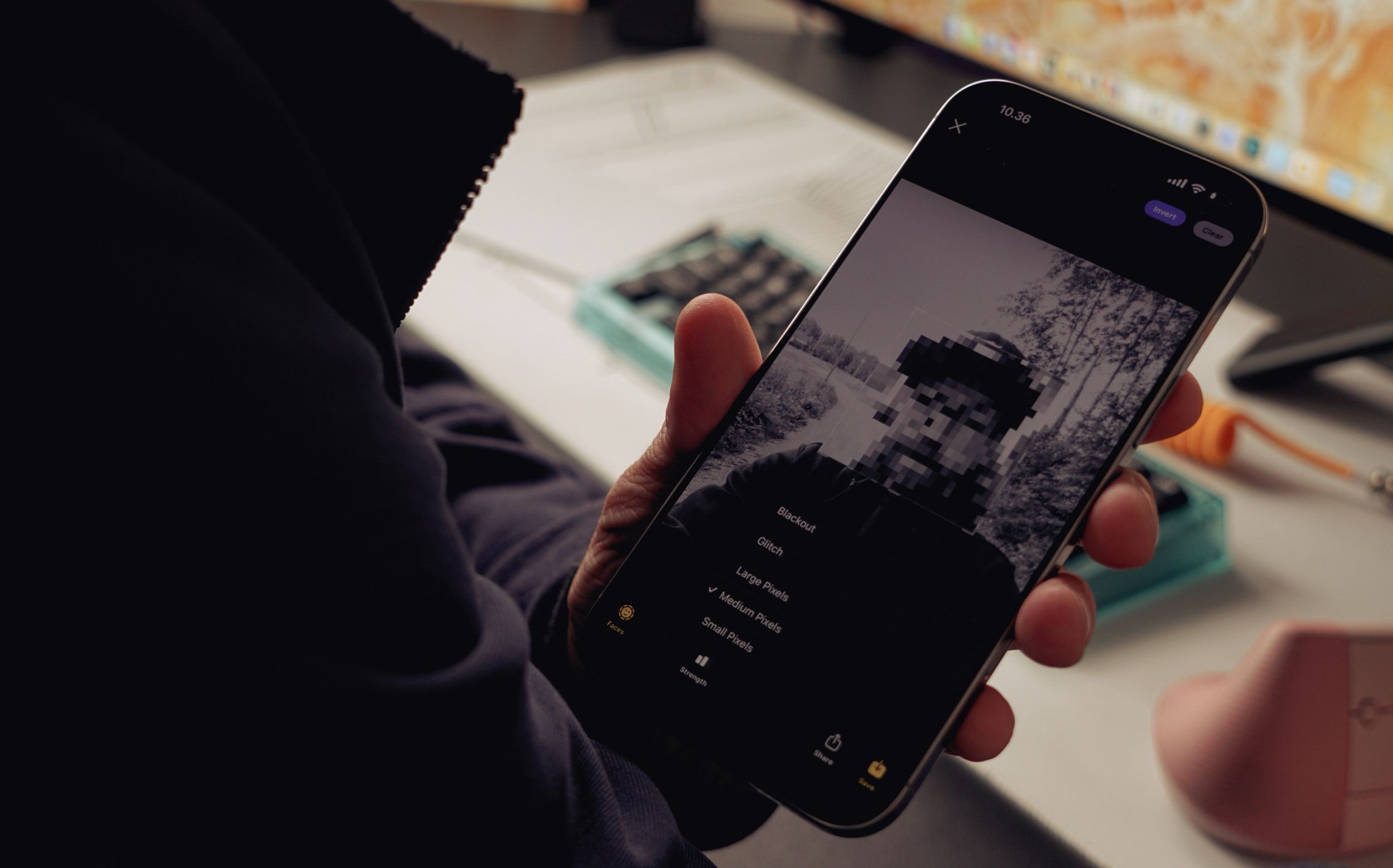 Das Bild zeigt eine Person, die ein Smartphone hält und eine bearbeitete Schwarz-Weiß-Fotoansicht betrachtet. Die Darstellung auf dem Bildschirm ist unscharf und pixelig. Im Hintergrund sind Gegenstände wie ein Computer und verschiedene Büroartikel zu sehen.