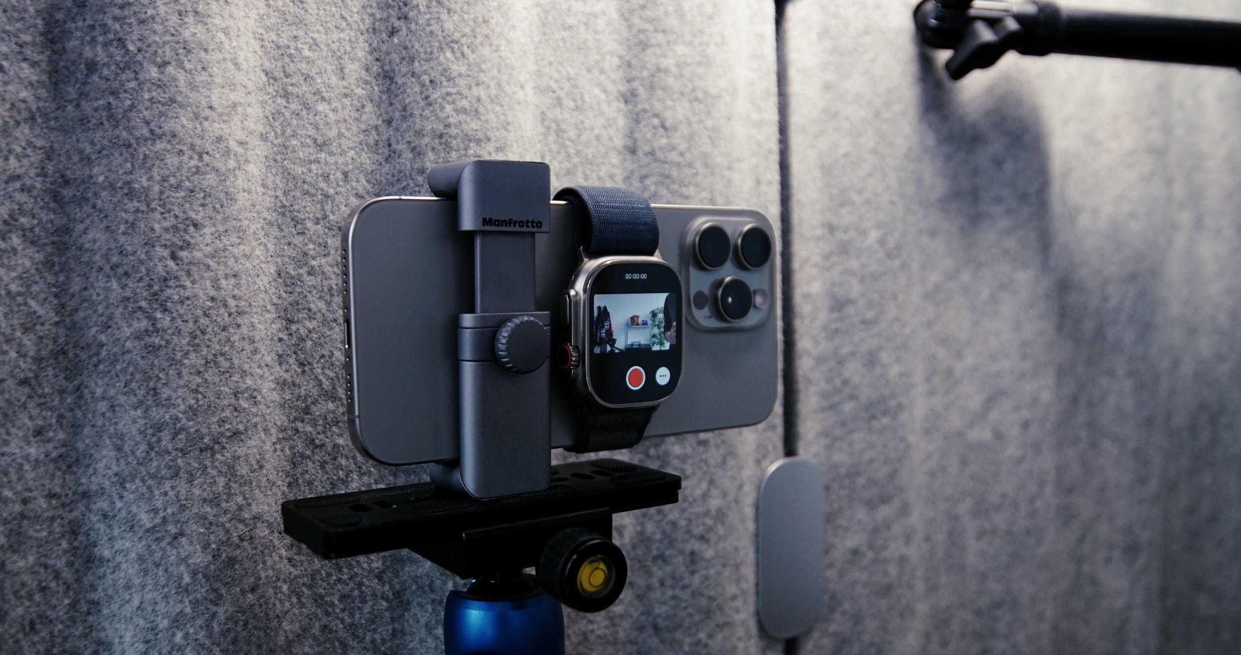 Das Bild zeigt ein Smartphone, das auf einem Stativ montiert ist, zusammen mit einer Smartwatch, die offenbar zur Aufnahme verwendet wird. Im Hintergrund ist eine graue Wand zu sehen, die aus einem strukturierten Material besteht. Diese Anordnung deutet auf eine professionelle Videoaufnahme oder Live-Streaming-Situation hin.