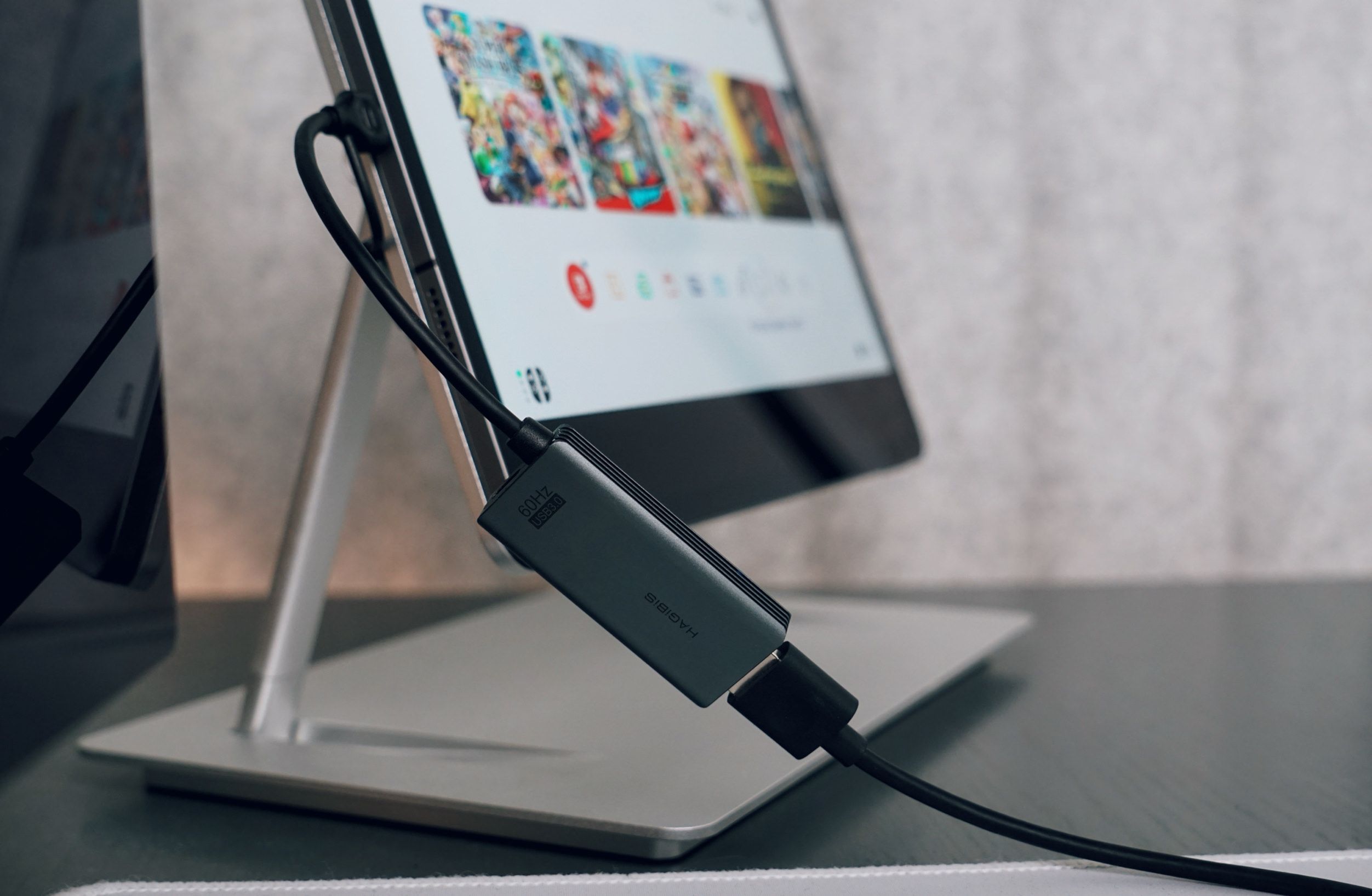 Close-up of a tablet displaying a gaming interface, connected by a black cable to a gray Hagibis USB 3.0 HDMI adapter. The tablet is propped on a silver stand on a dark surface.