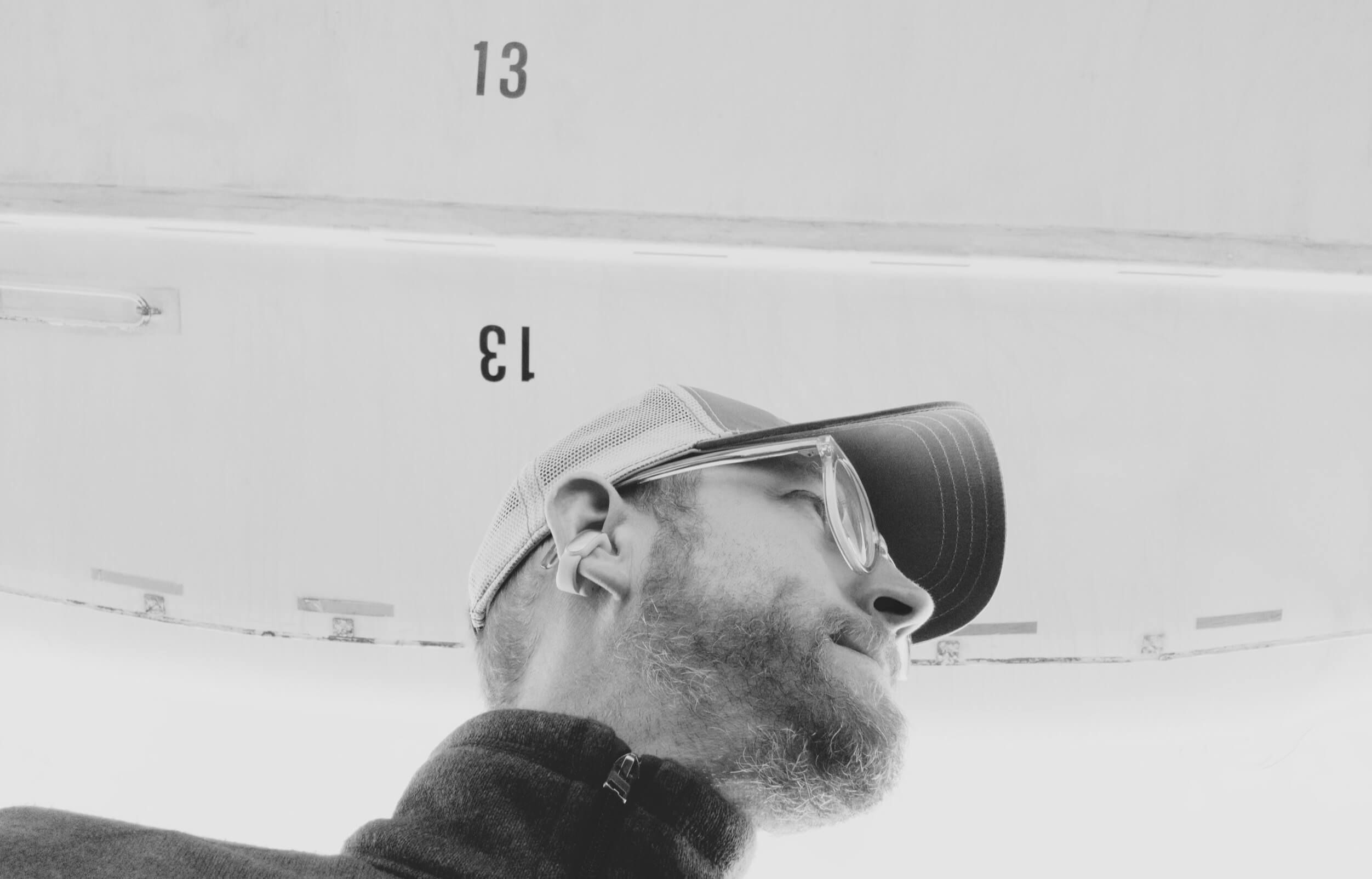 Black and white photo of a bearded man looking up towards a plain white surface bearing the number "13". The man wears a baseball cap and glasses. The photo is angled upwards.
