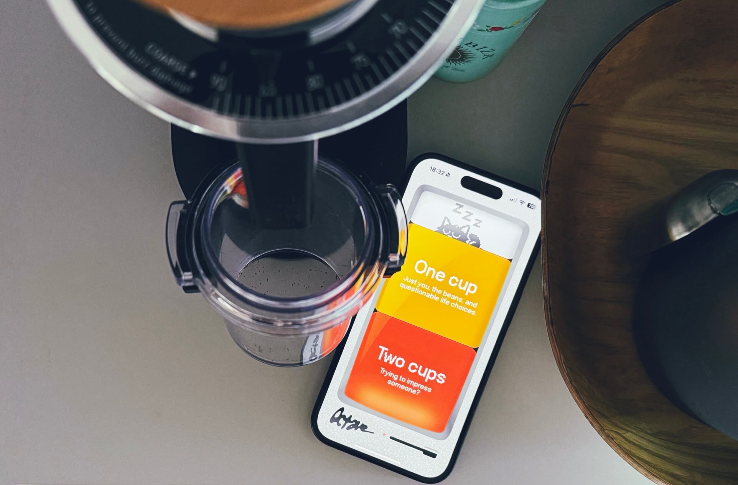 Overhead shot of a coffee grinder with a clear container below it, alongside a smartphone displaying an app for selecting coffee cup amounts, with the choices "One cup" and "Two cups" and associated descriptions.