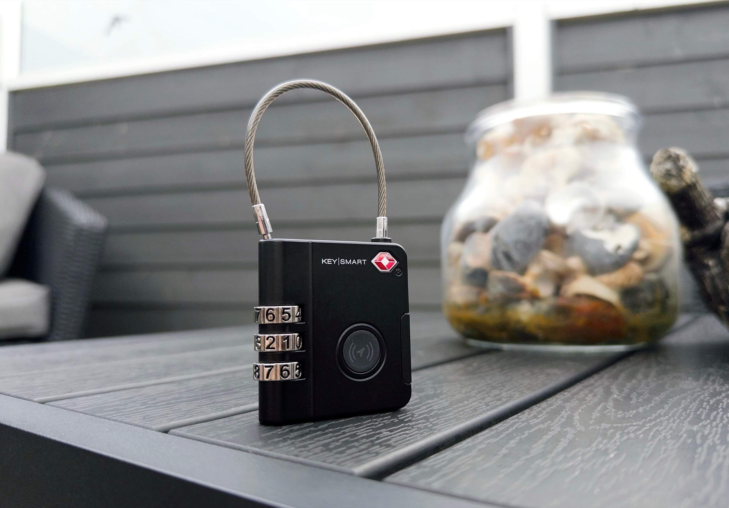 A black KeySmart combination lock with a silver cable, set on a grey wooden table, with a jar of rocks and seashells in the background.