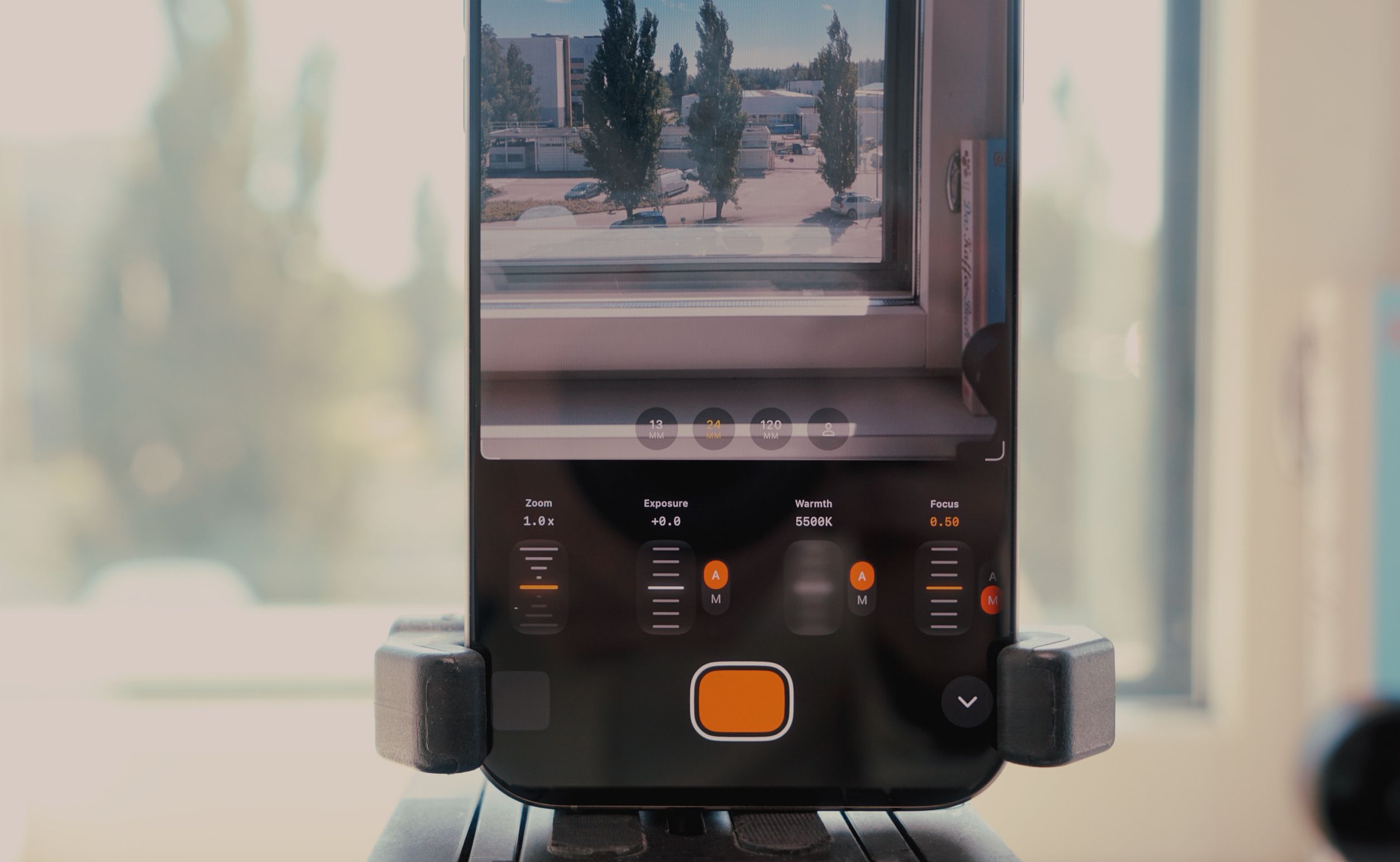 Smartphone screen displaying a live camera feed of a window with trees, and adjustment tools for zoom, exposure, warmth, and focus, mounted on a stand.