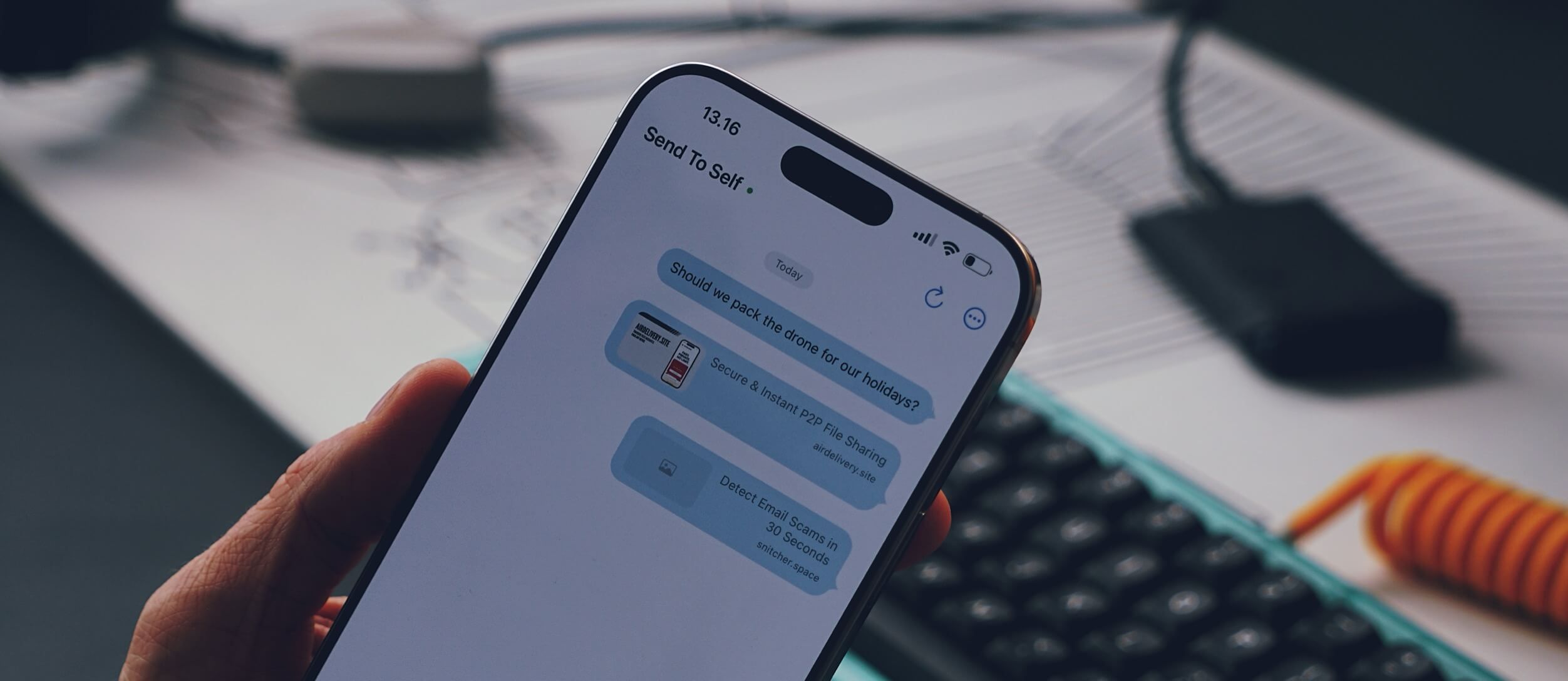 A hand holding a smartphone displaying a messaging app conversation with "Send To Self." The conversation includes three blue message bubbles: "Should we pack the drone for our holidays?," "Secure & Instant P2P File Sharing, airdelivery.site," and "Detect Email Scams in 30 Seconds, snitcher.space." The time 13:16 is shown at the top, along with the phone's status bar. The background shows a partially visible desk, a keyboard, and some cables.