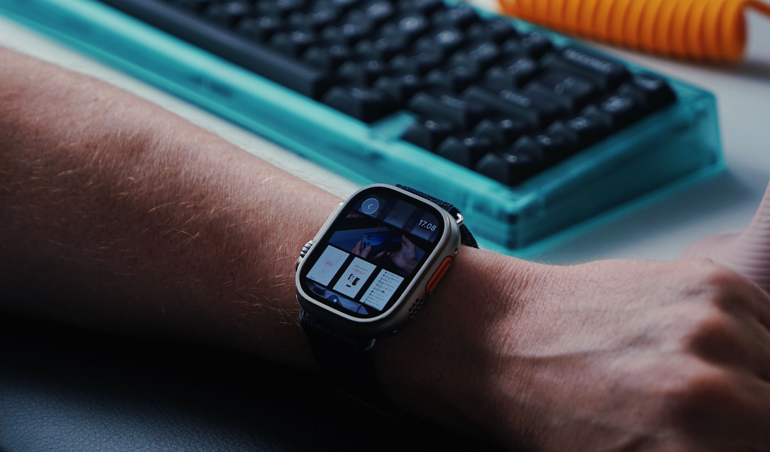 Close-up shot of a wrist with a silver smartwatch displaying a user interface, likely showcasing app information or a video, with a blue strap. The wrist is resting near a clear turquoise mechanical keyboard, and an orange cable. The watch face shows the time, 17:08.
