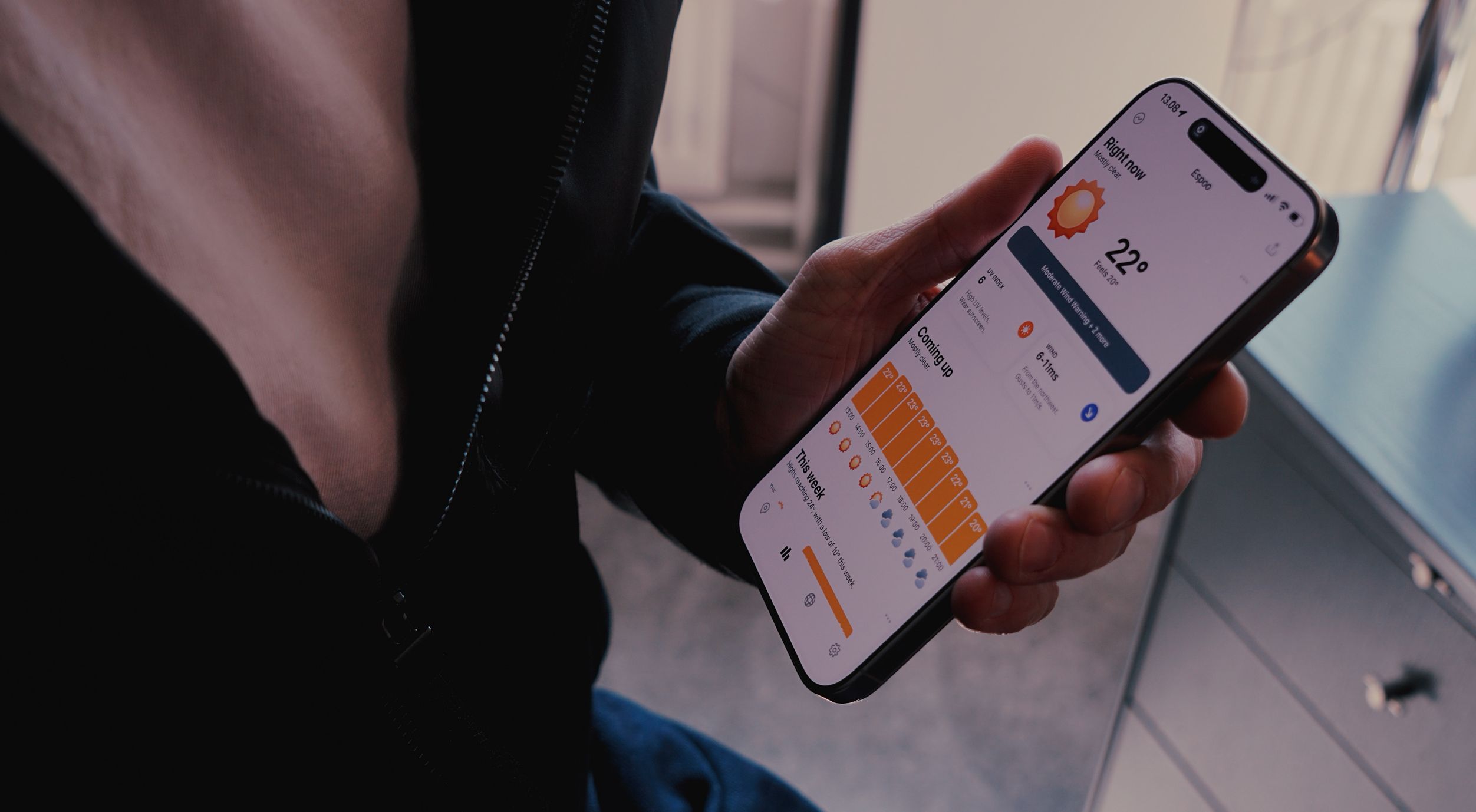 A person's hand holding a smartphone displaying a weather forecast for Espoo. The screen shows a sunny icon, a temperature of 22 degrees Celsius, and a 'Moderate Wind Warning'. The weather forecast includes 'Coming up' hourly details and a 'This week' overview. The person is wearing a dark jacket, with the zipper in focus.