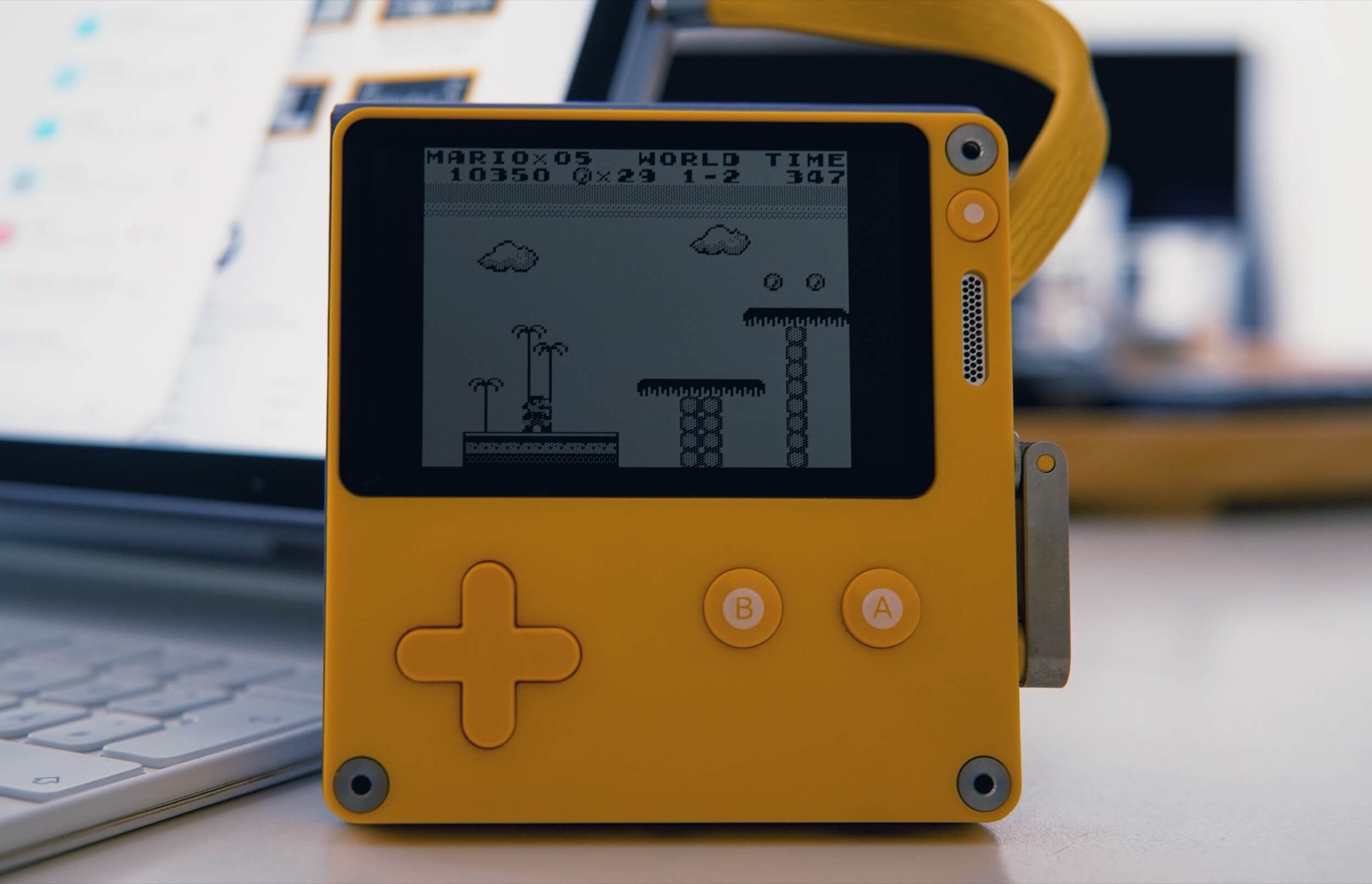 A yellow handheld gaming console displaying a black and white image of Super Mario Bros gameplay, with a yellow cross-shaped directional pad, "B" and "A" buttons, and a connecting strap visible, placed on a white surface with a partially visible laptop in the background.
