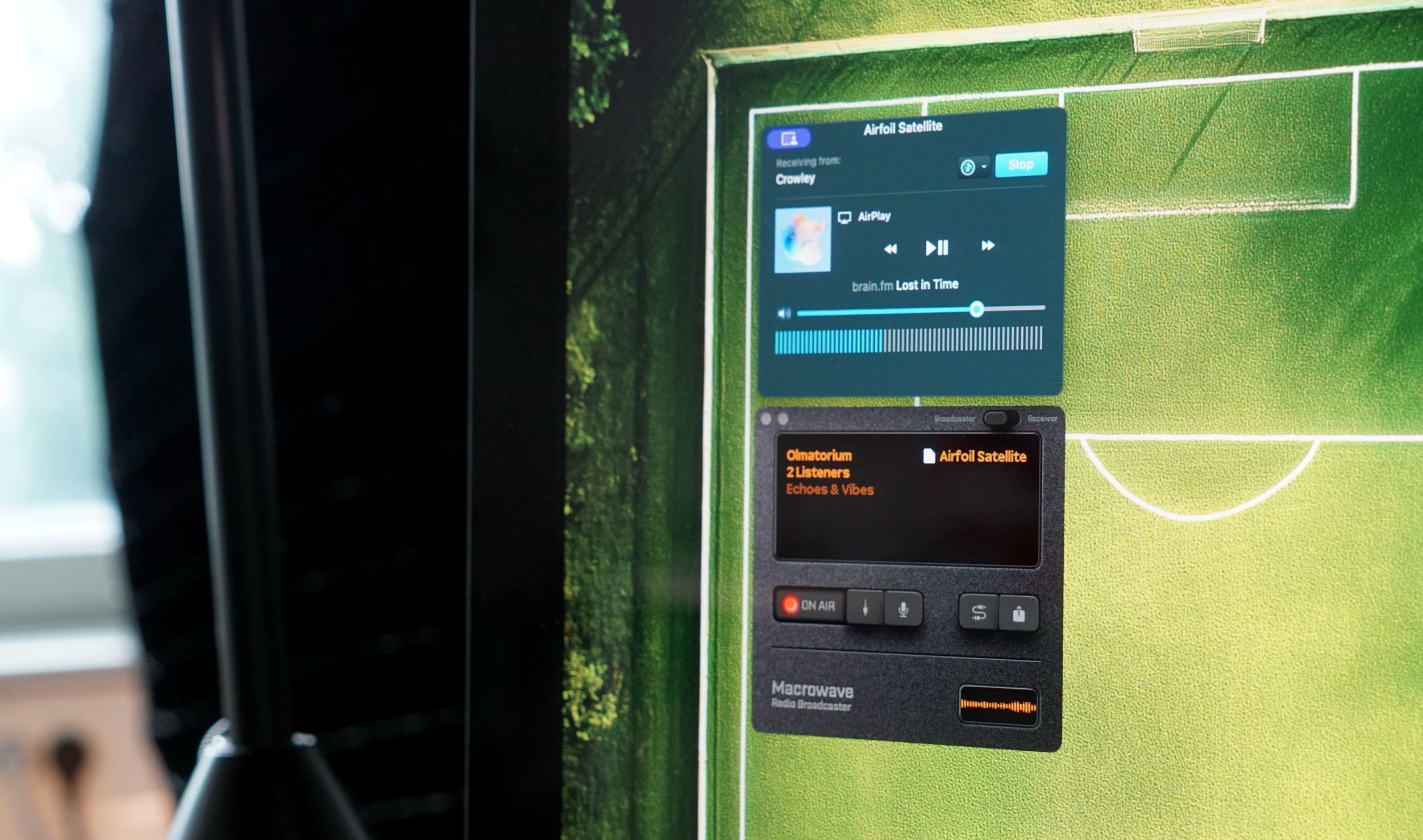 Close-up of a computer monitor displaying a music player and broadcast control panel overlaid on a soccer field graphic. The music player, labeled "Airfoil Satellite," shows "brain.fm - Lost in Time" playing via AirPlay, receiving from "Crowley." Below, a "Macrowave" radio broadcaster control panel indicates "ON AIR" status, with additional controls visible.
