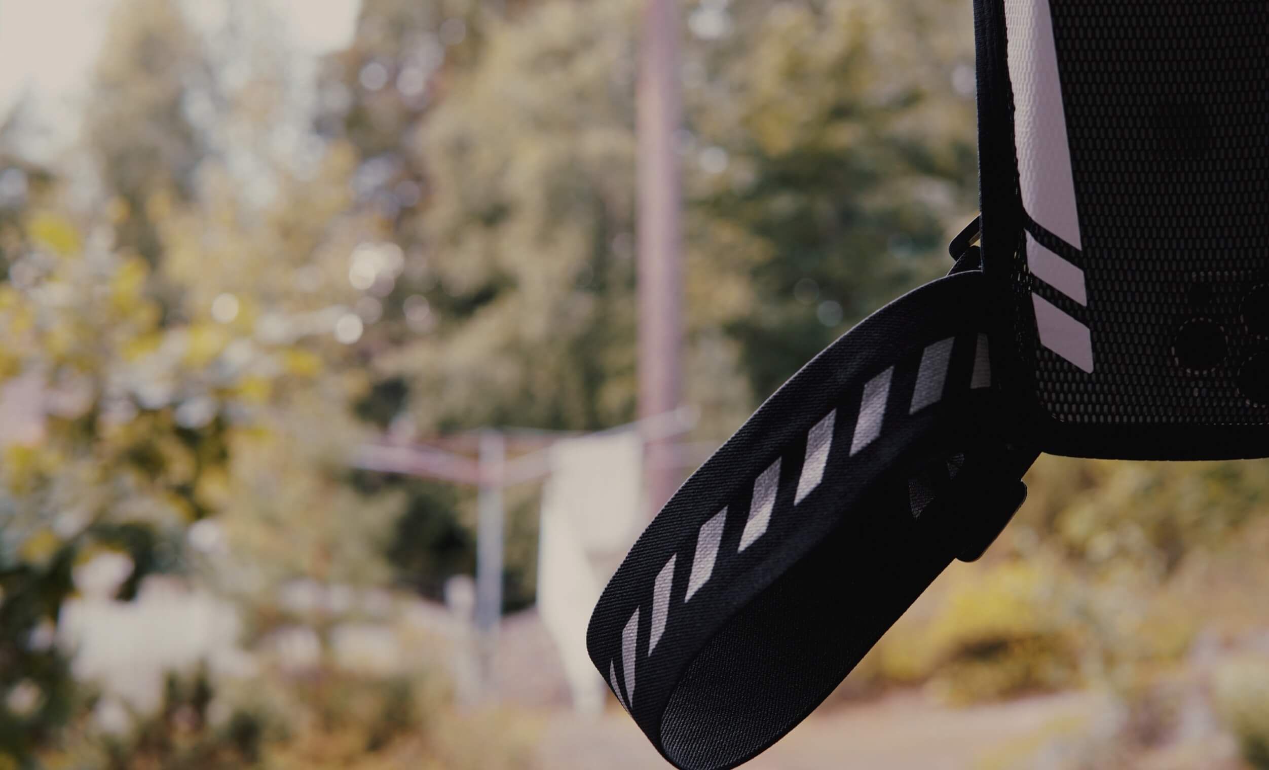Close-up shot of a black backpack strap, featuring reflective white diagonal stripes, against a blurred, natural outdoor background. The strap appears to be made of a durable, textured material.

